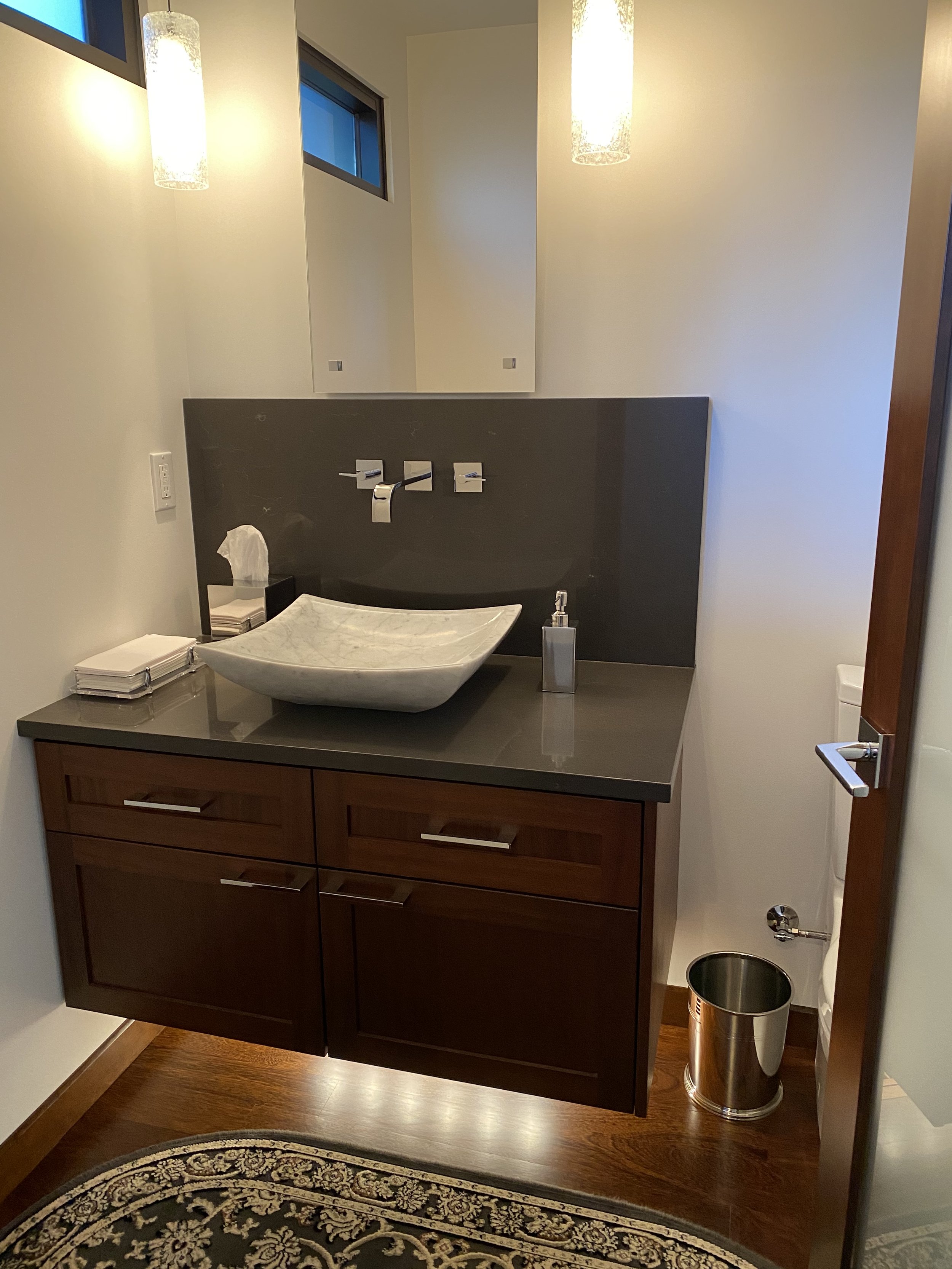 Modern bathroom vanity with a white vessel sink, a dark countertop, wooden cabinets, and a soap dispenser. Wall-mounted faucet above the sink, with mirror and window in the background. Tissues and towels on the counter. Small trash can near the toile