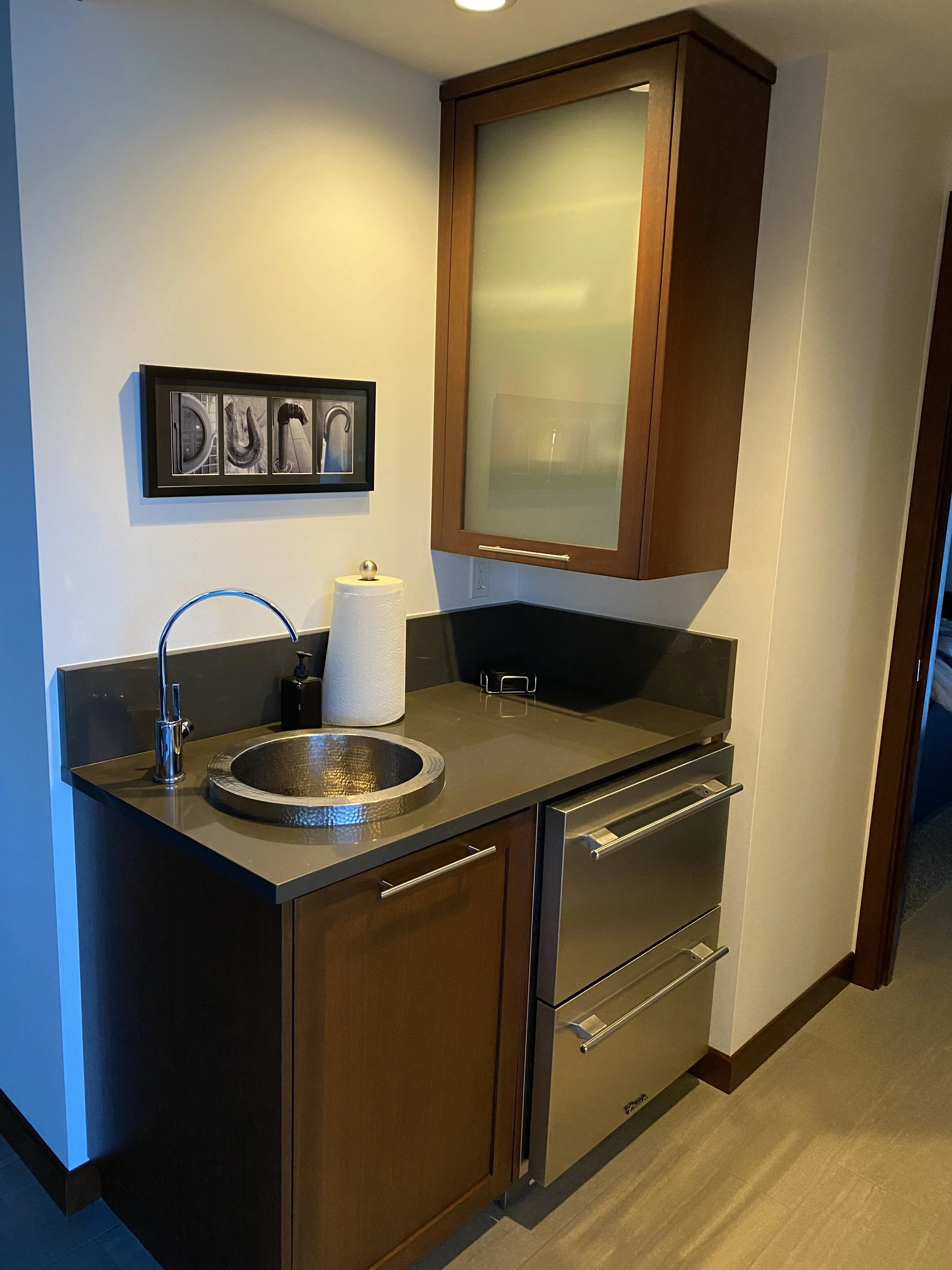A small kitchen area with a wooden cabinet above a black countertop, a stainless steel sink with a curved faucet, a paper towel holder, a soap dispenser, a microwave oven, and a framed black-and-white artwork on the wall.
