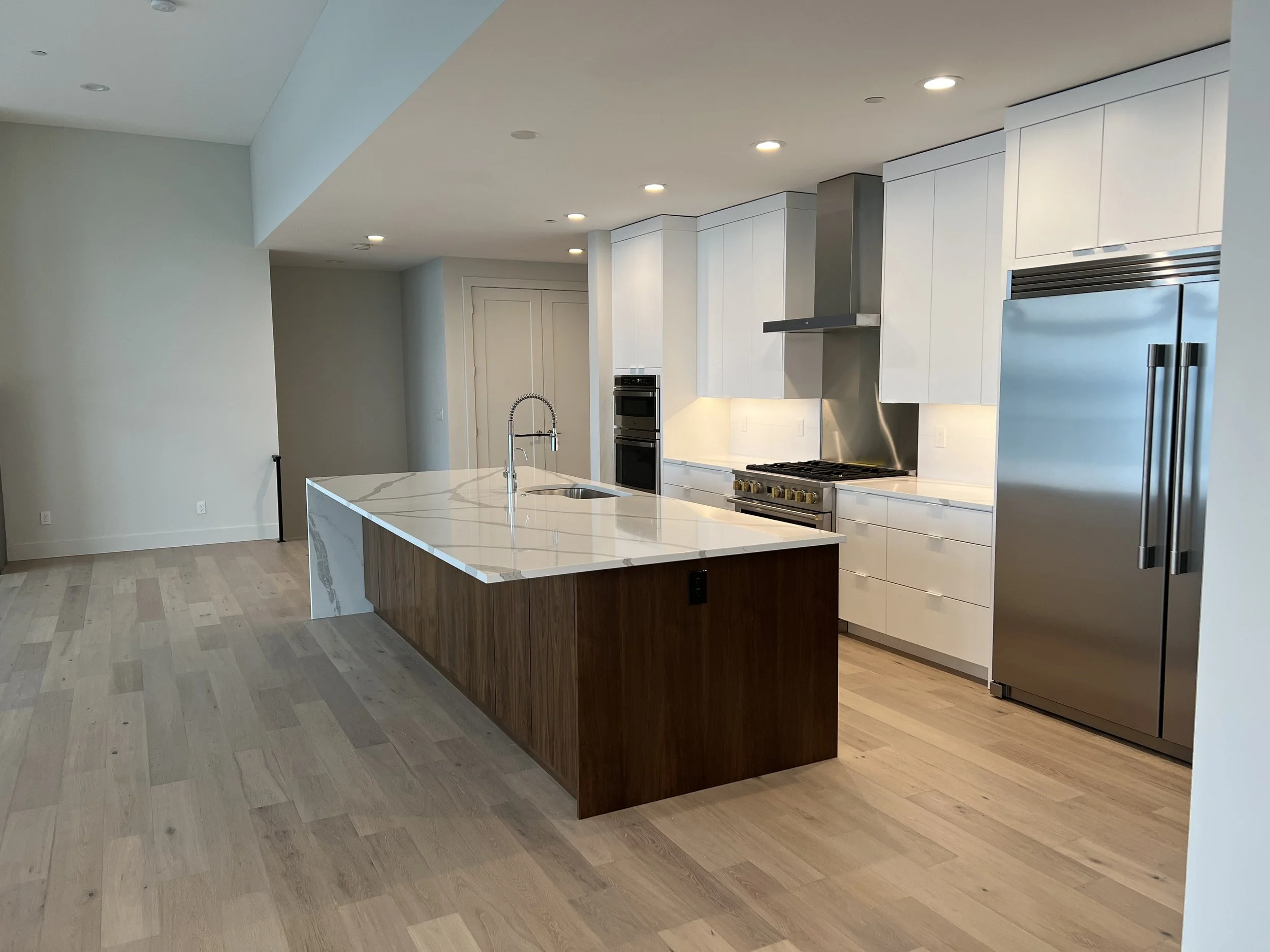 Modern kitchen with white cabinetry, stainless steel appliances, a large island with a marble countertop, and light-colored hardwood floors.