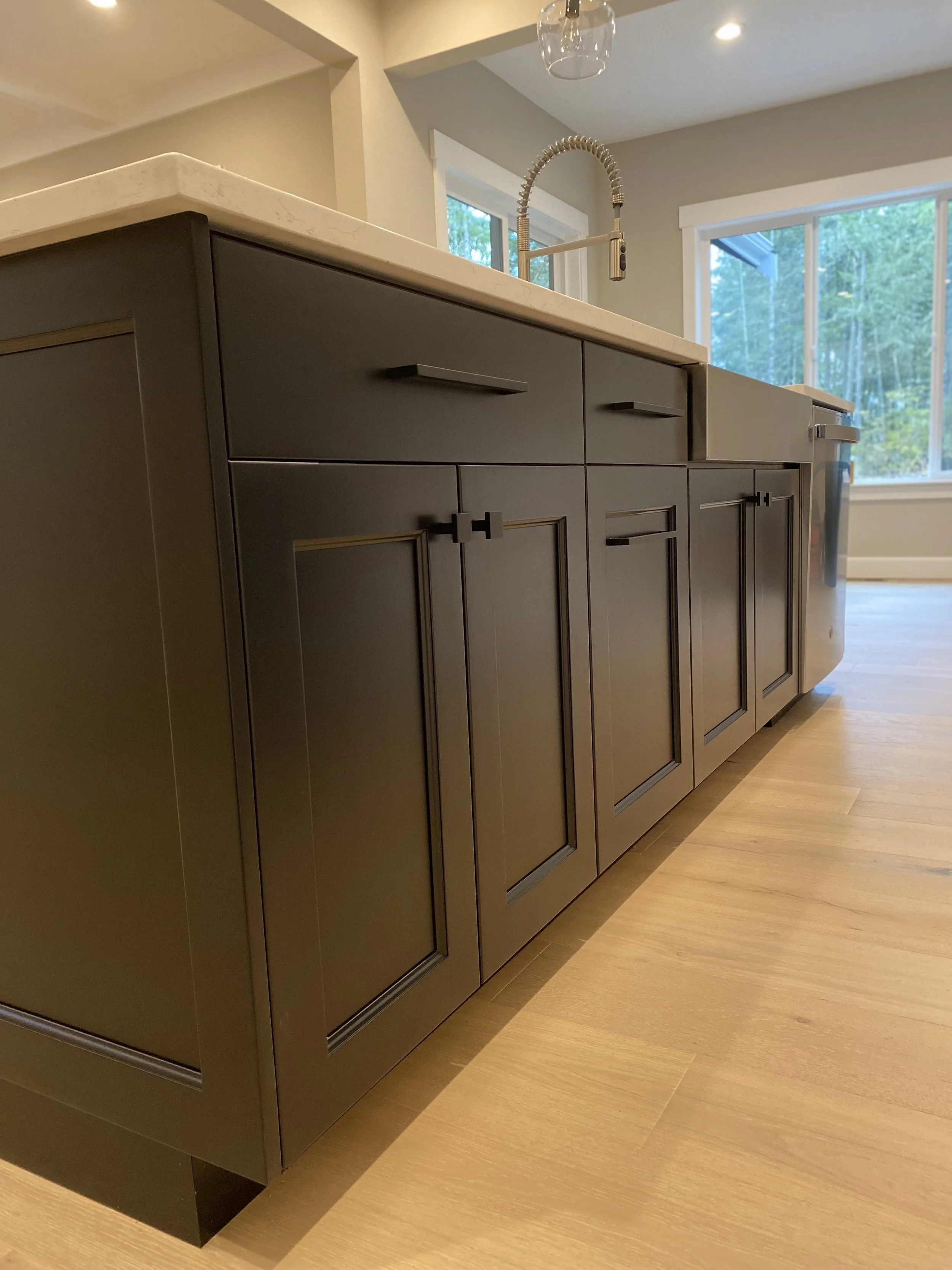 Close-up of a kitchen island with a beige countertop, dark cabinetry with paneled doors, and a gold-colored handle. The faucet is metallic with a flexible spring neck. In the background, large windows reveal a view of trees outside.