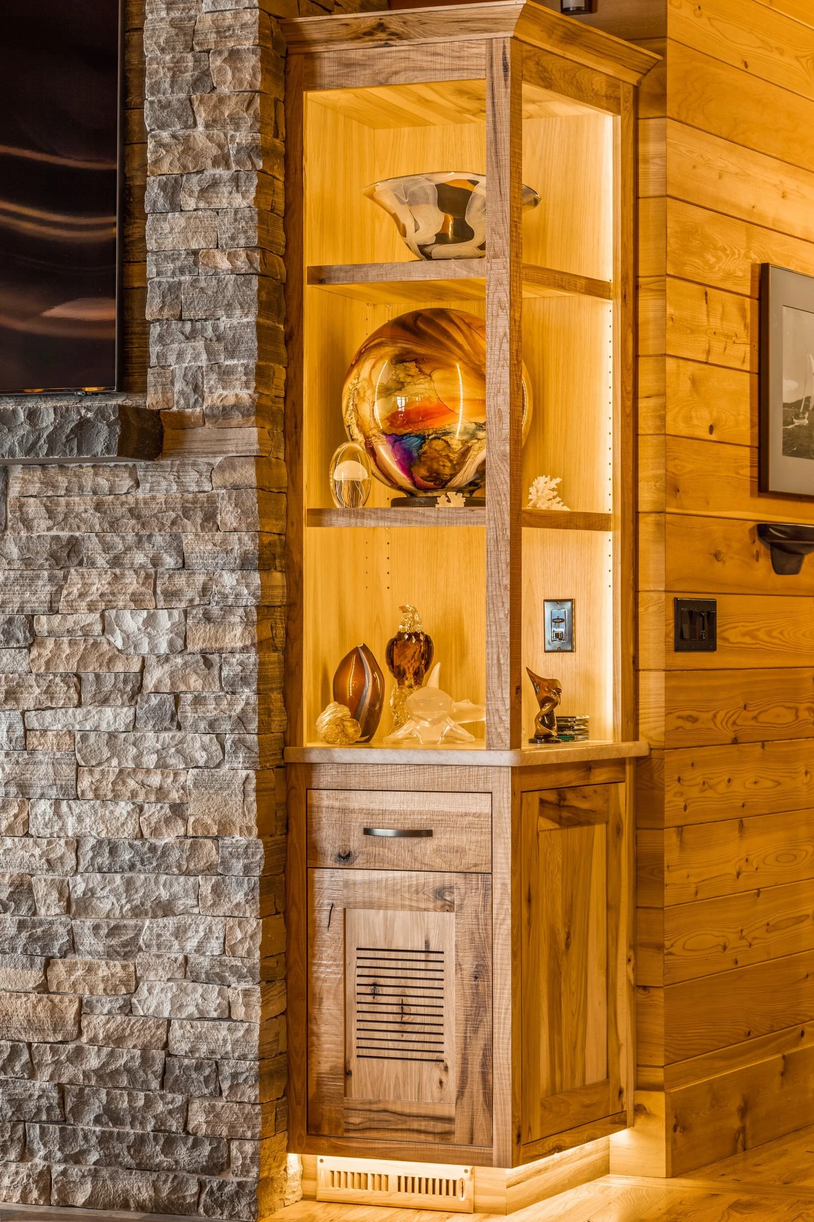 Decorative glass sculptures displayed on wooden shelves built into a corner of a rustic room, with a stone wall on the left and wood-paneled wall on the right.