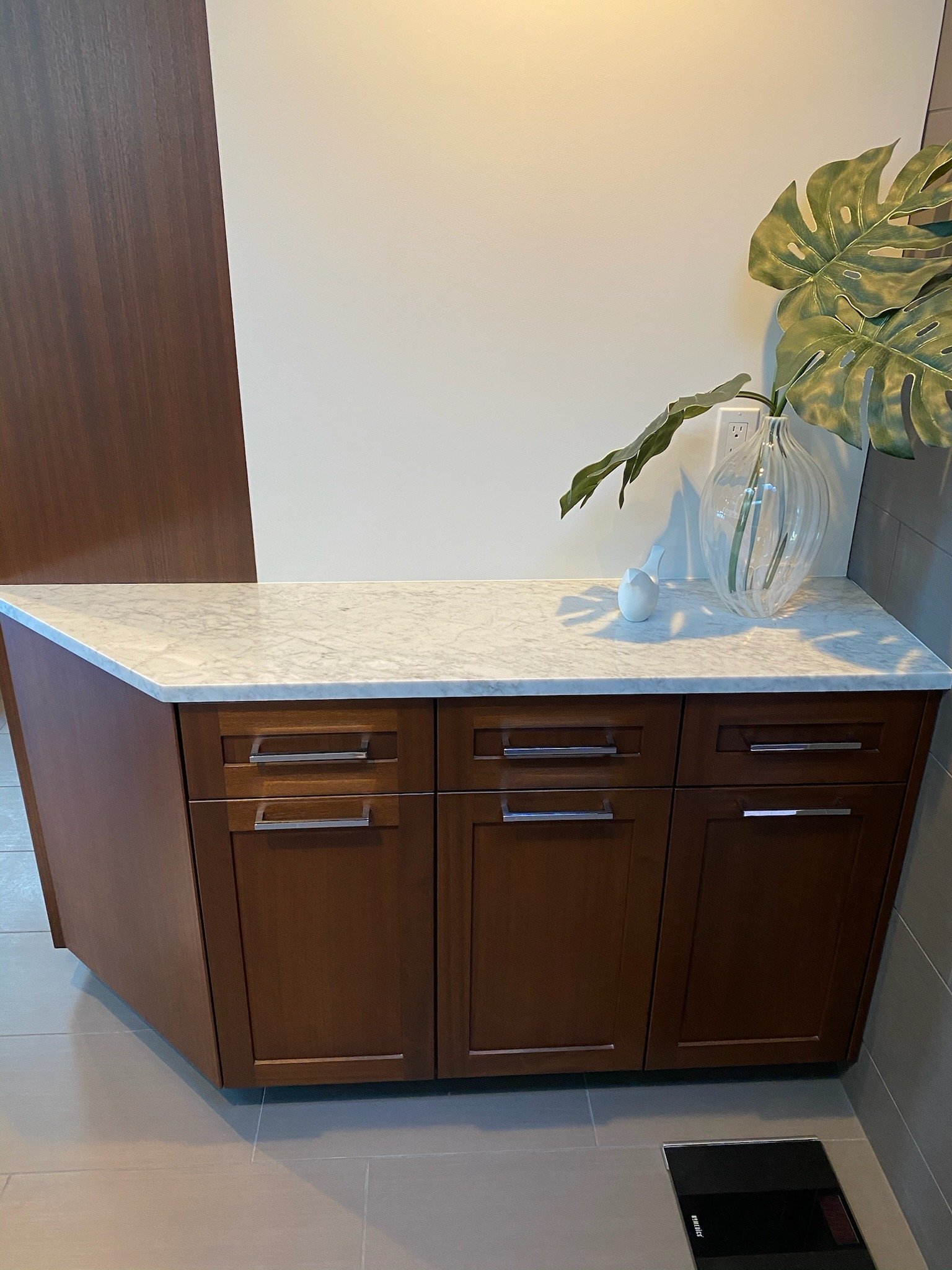 A wooden sideboard with a marble top, decorated with a large glass vase containing green leaves and a small white decorative object, set against a plain wall and tiled floor.