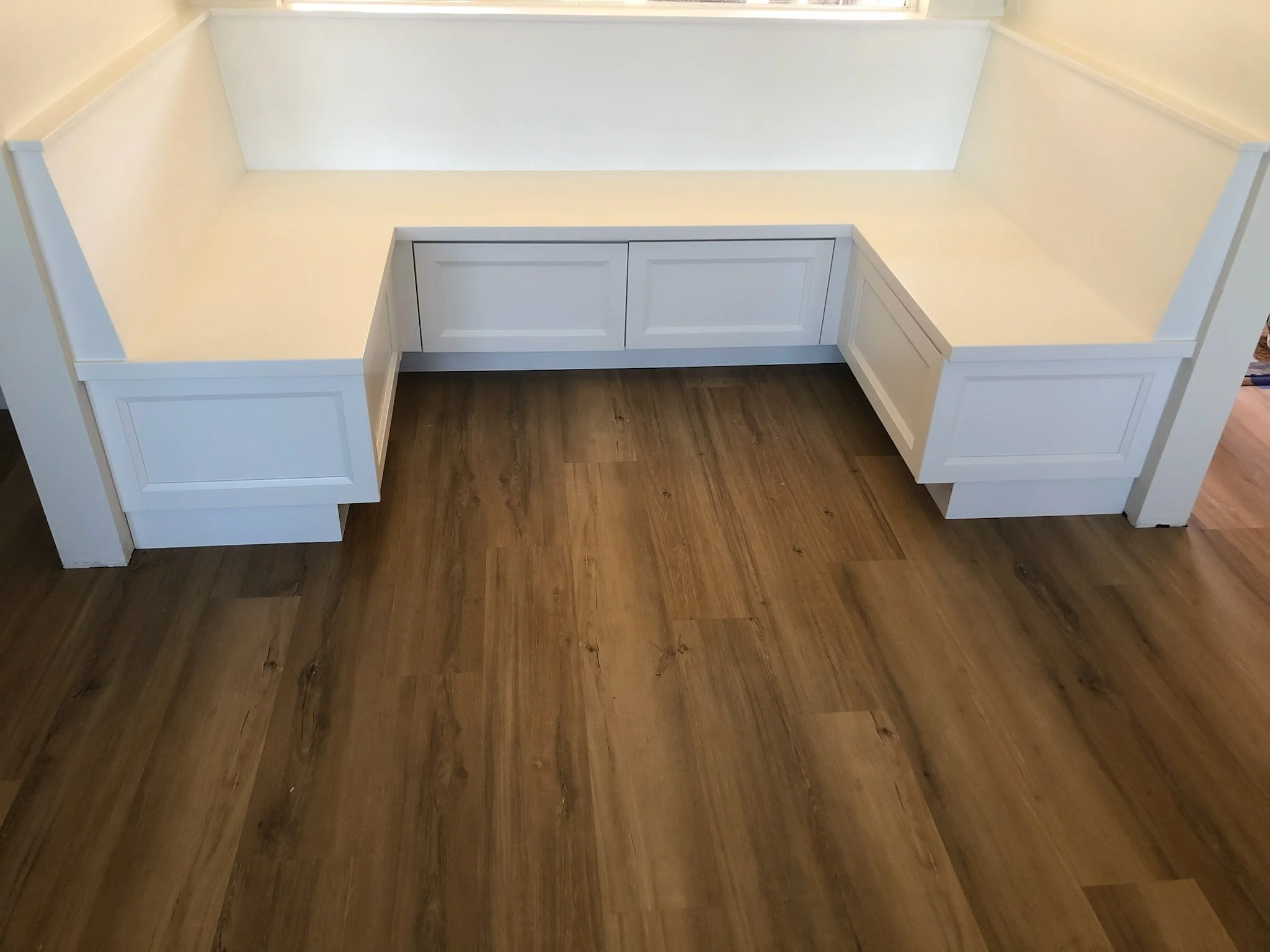 White built-in bench with storage drawers and cushions, positioned against a wall with hardwood flooring.