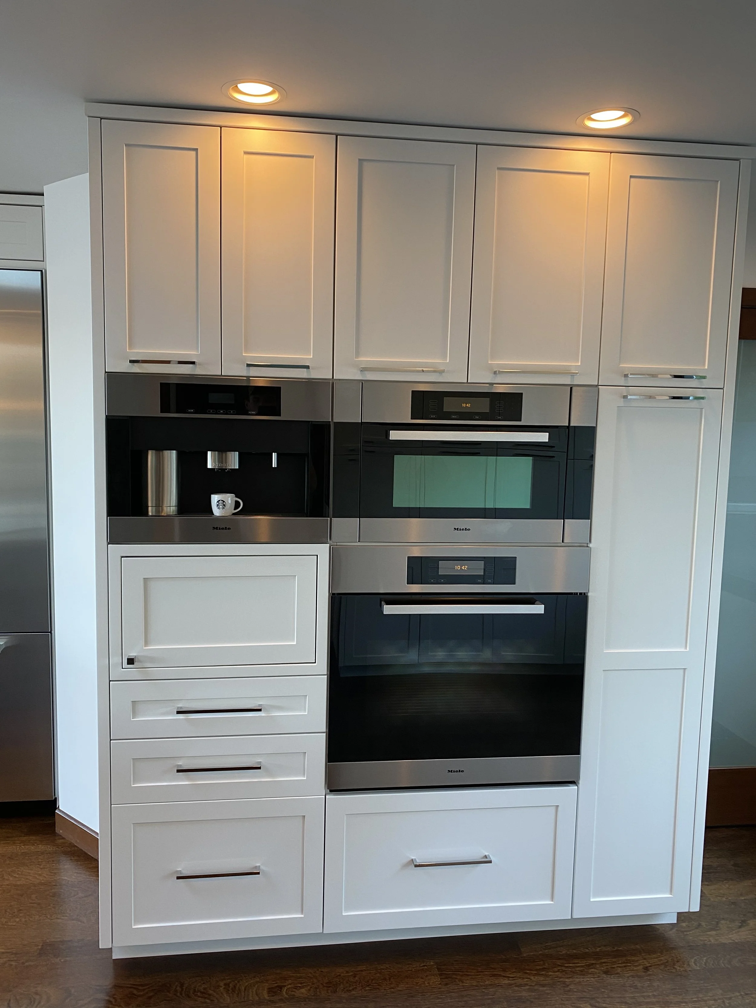 Kitchen cabinetry with built-in coffee maker, oven, microwave, and storage drawers, illuminated by recessed ceiling lights.