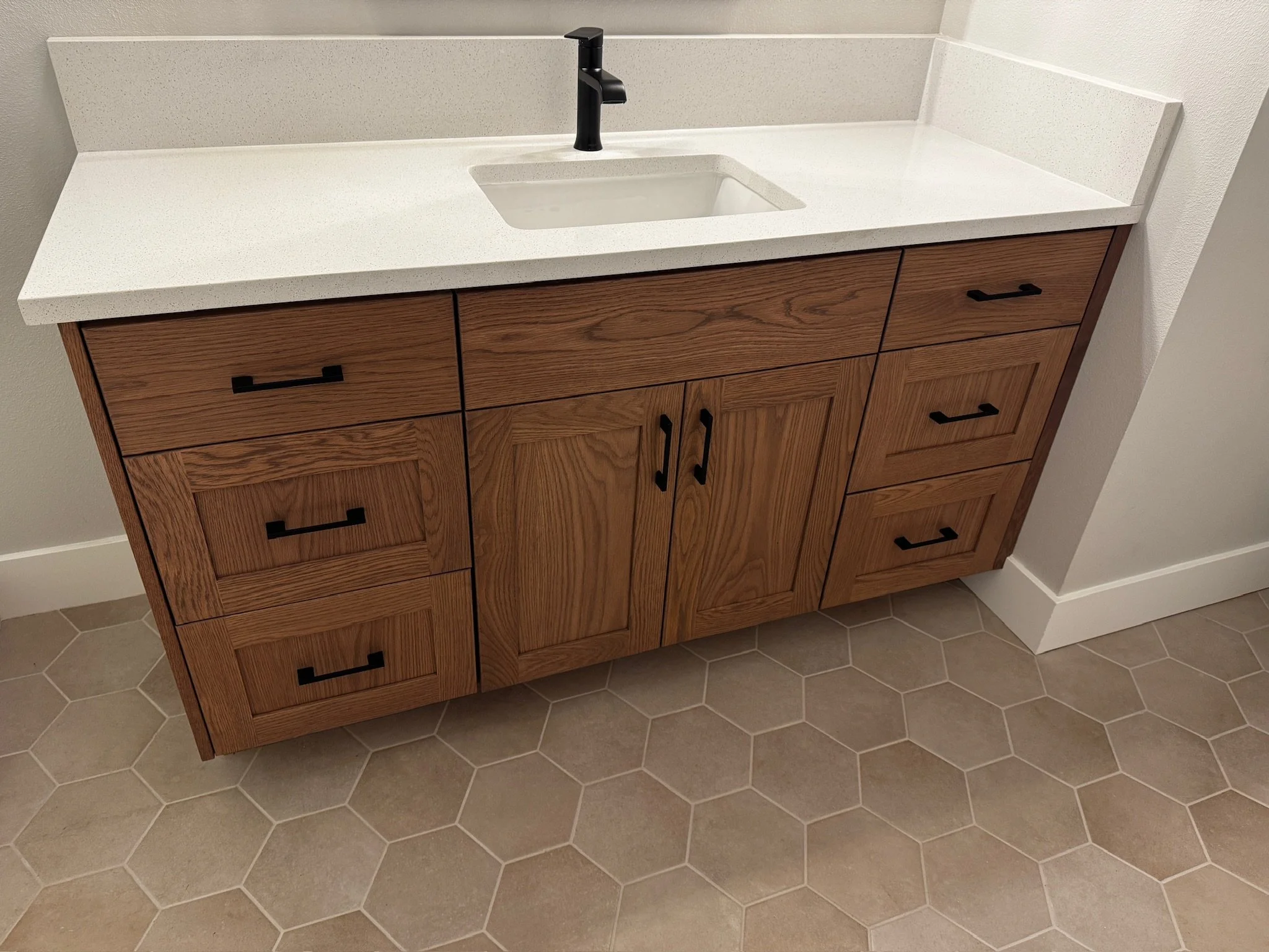 Wooden bathroom vanity with a white countertop, black handles, and a black faucet, against a white wall and hexagonal tile floor.