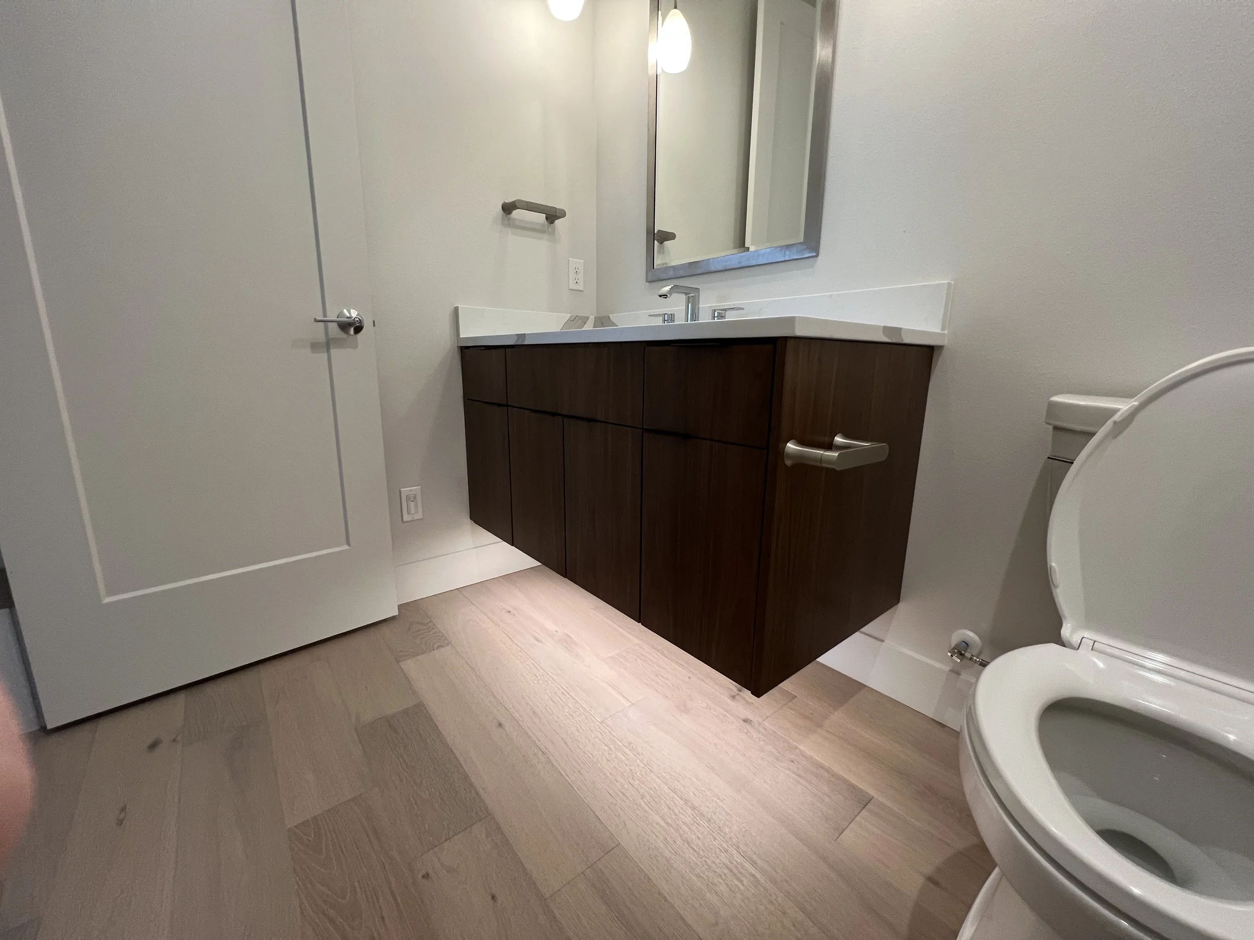 Modern bathroom with wooden vanity, mirror, white sink, towel rack, toilet, and wood flooring.