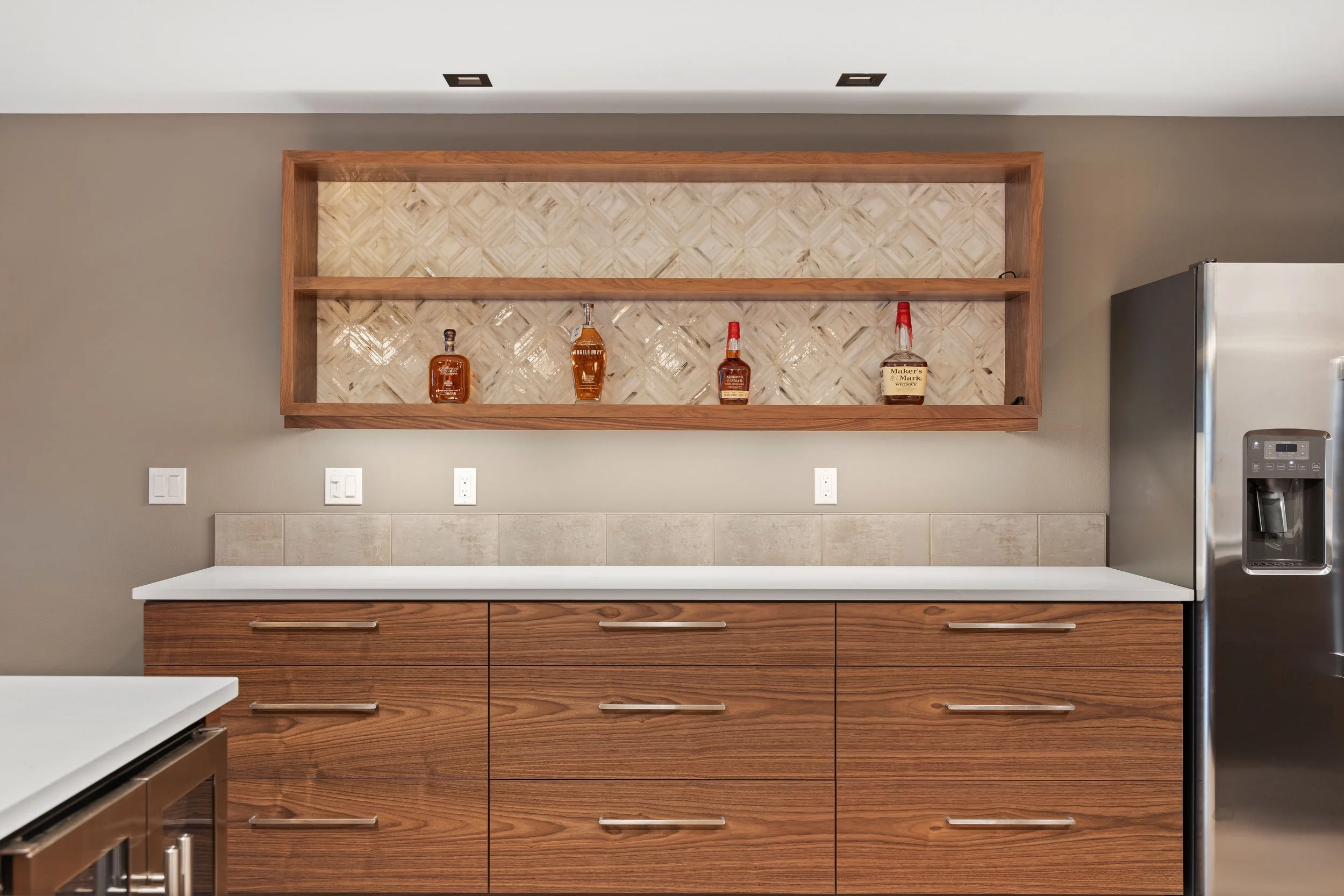 A modern kitchen with wooden cabinets, a white countertop, a beige tile backsplash, and a wooden open shelf above displaying four bottles of liquor. A stainless steel refrigerator is on the right.