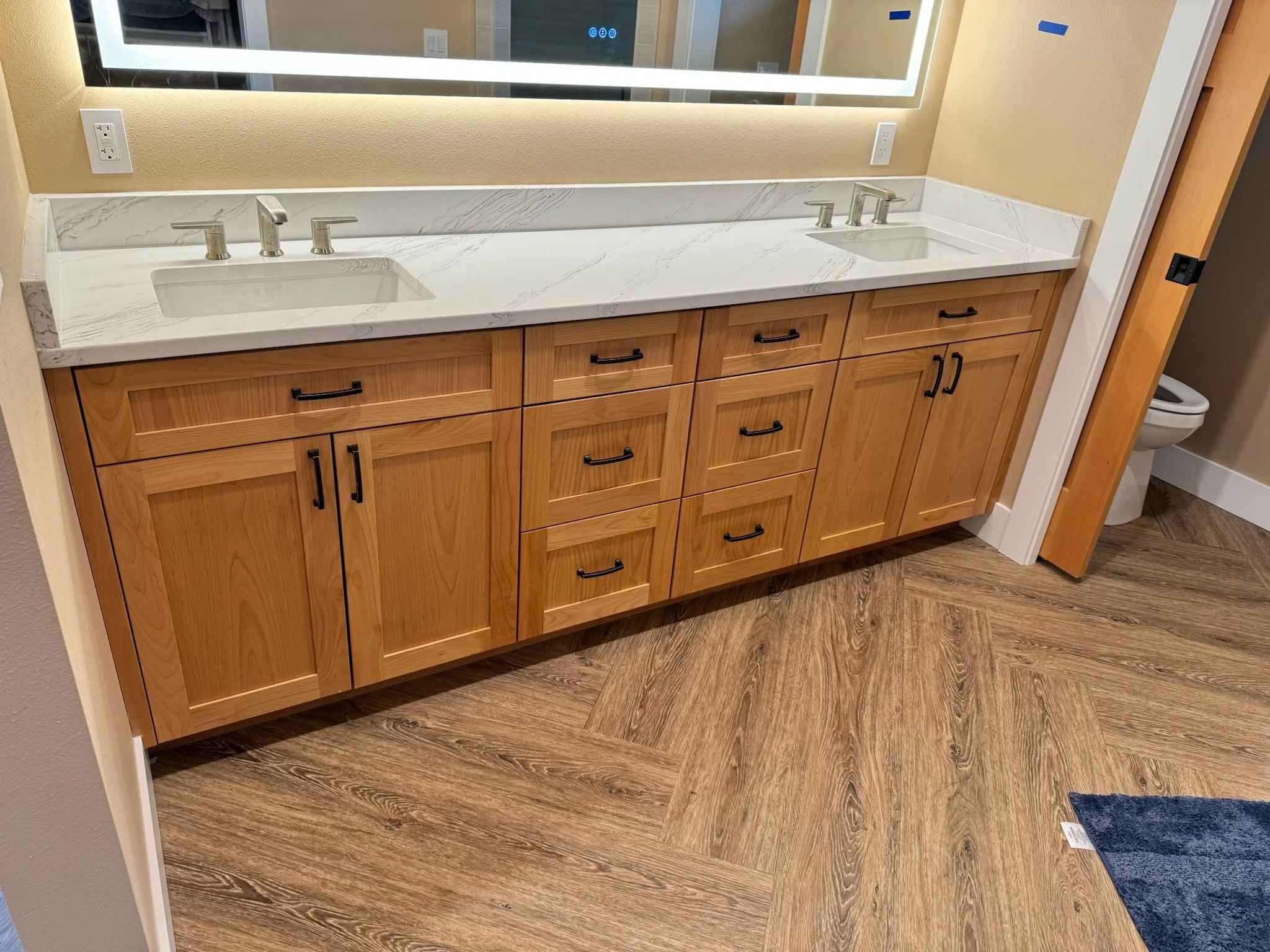 Bathroom vanity with double sinks, wooden cabinets, and a marble countertop, with a mirror above and a glimpse of a toilet in the background.