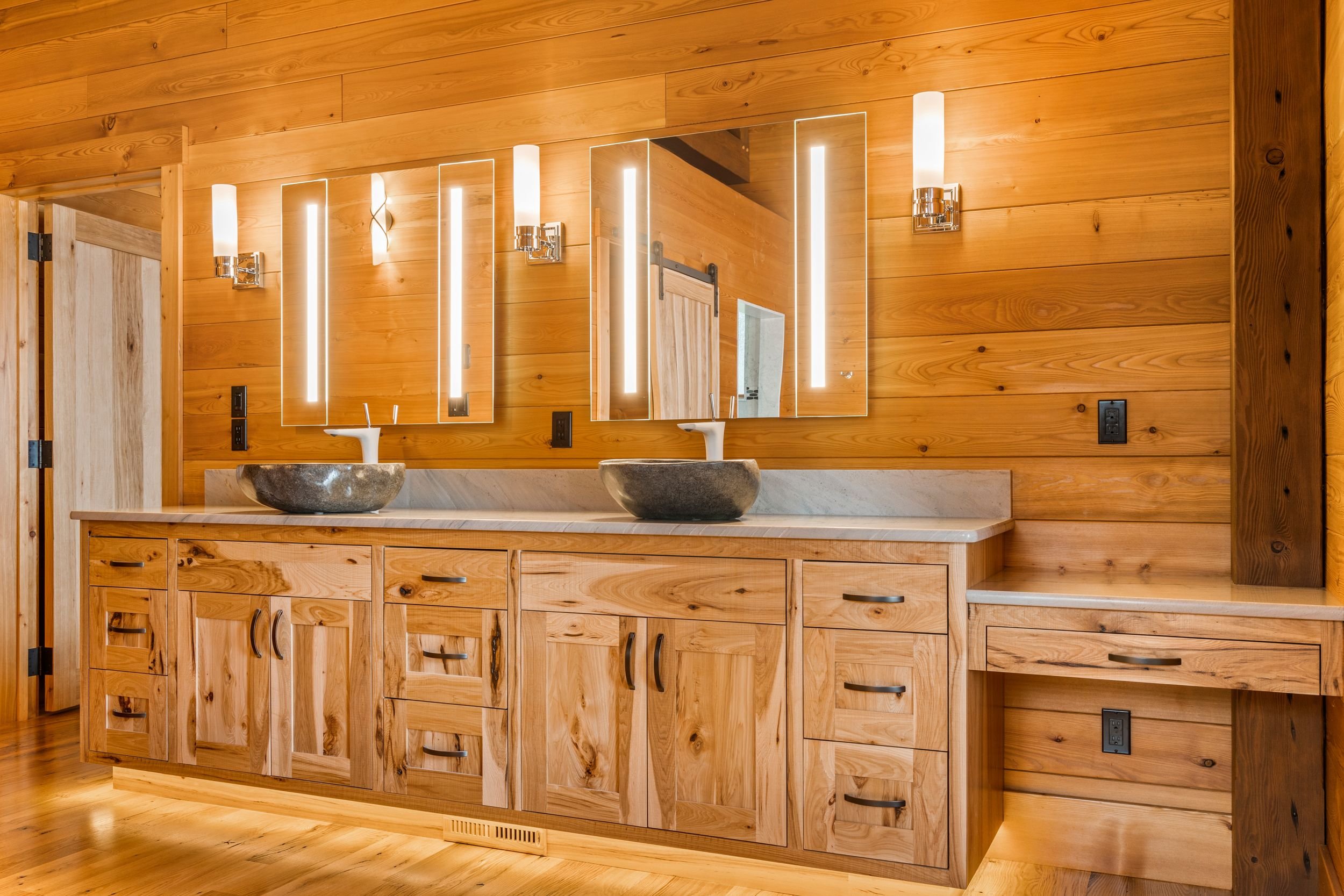 Bathroom with wood-paneled walls and cabinetry, two stone vessel sinks on a marble counter, illuminated mirrors, and modern wall-mounted light fixtures.