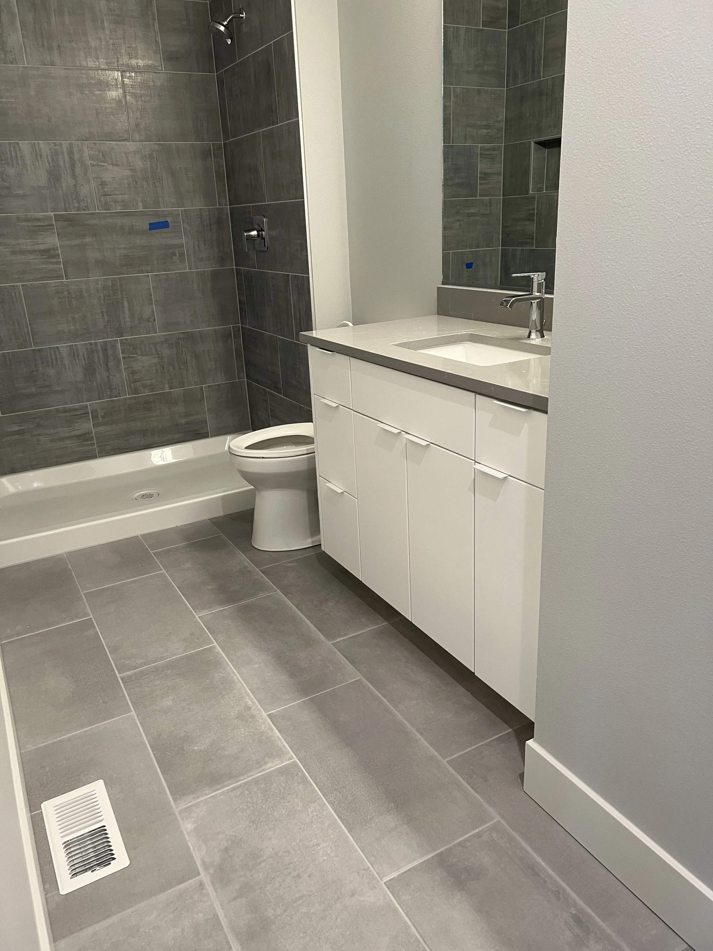 Modern bathroom with gray tiled shower and floor, white toilet, and white vanity with a sink and a mirror.