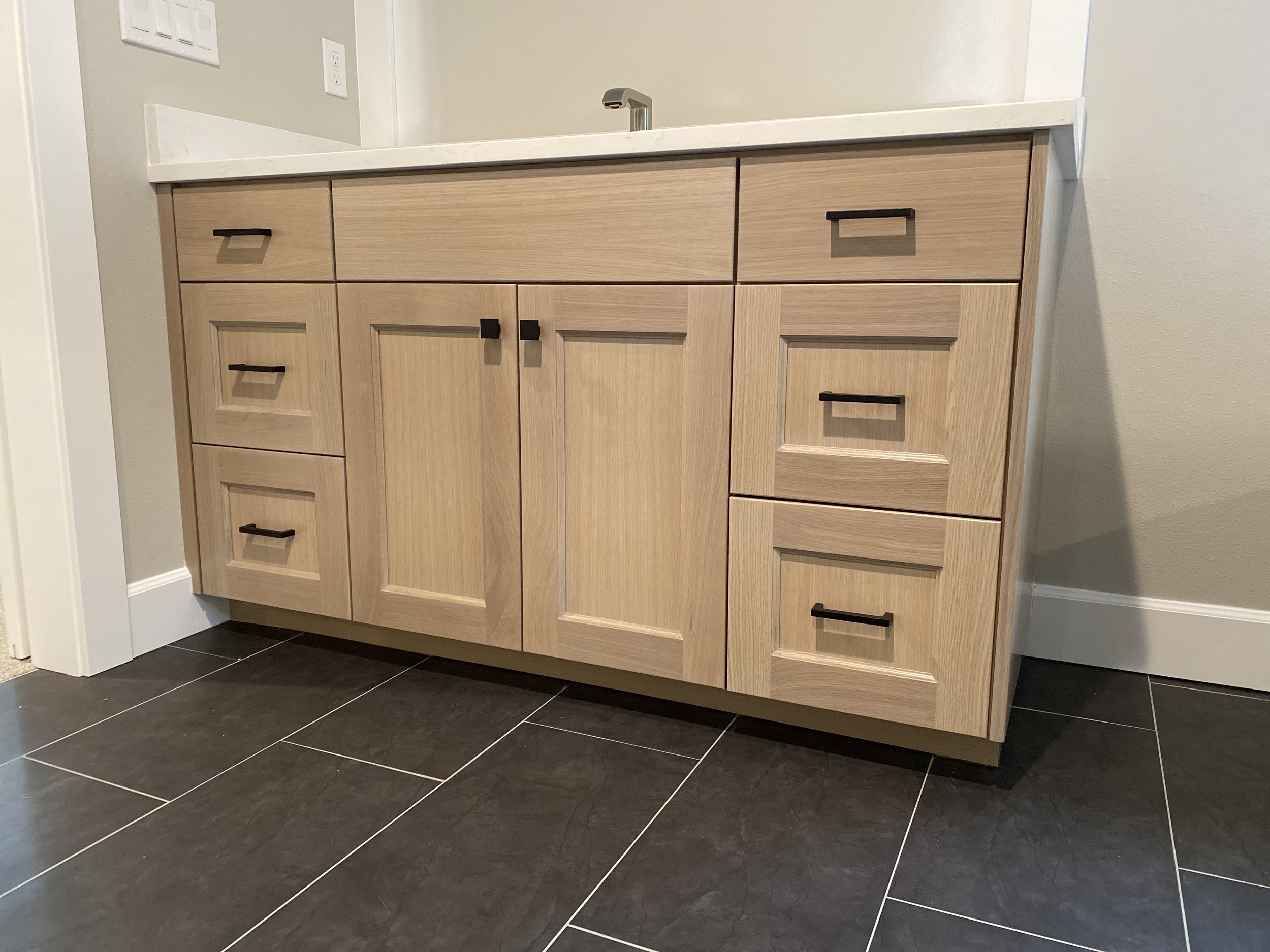 A modern bathroom vanity with light wood cabinets and black handles, a white marble countertop, and a silver faucet. The vanity is placed against a beige wall with a white baseboard, and the floor is covered with black tiles.