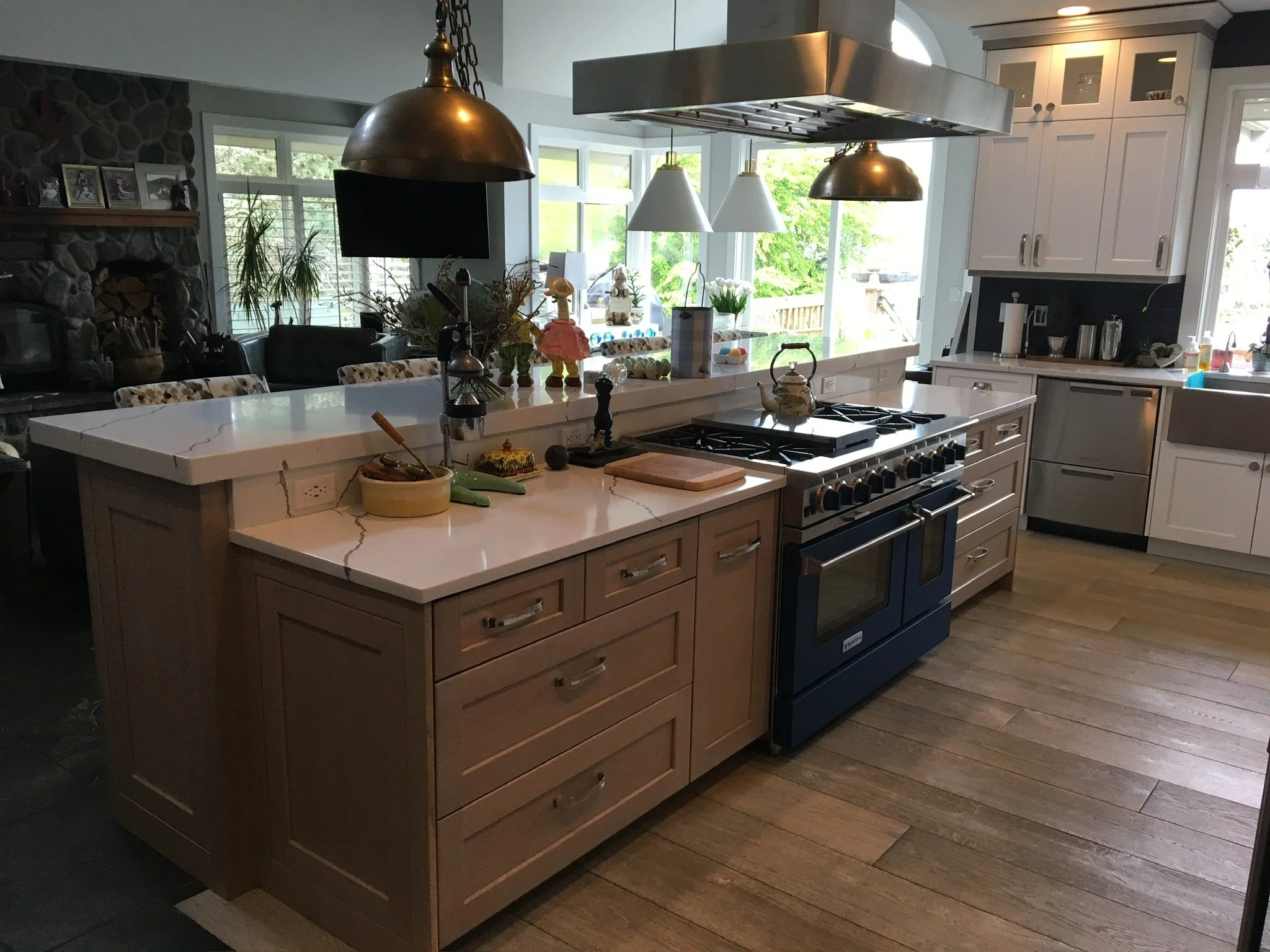 Modern kitchen with a central island, white and light wood cabinets, stainless steel appliances, pendant and track lighting, and large windows showing a green outdoor area.
