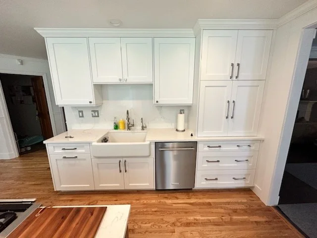 White kitchen cabinets with stainless steel dishwasher, wooden floor, and small countertop with a yellow and green soap dispenser, paper towel roll, and a sink with a faucet.