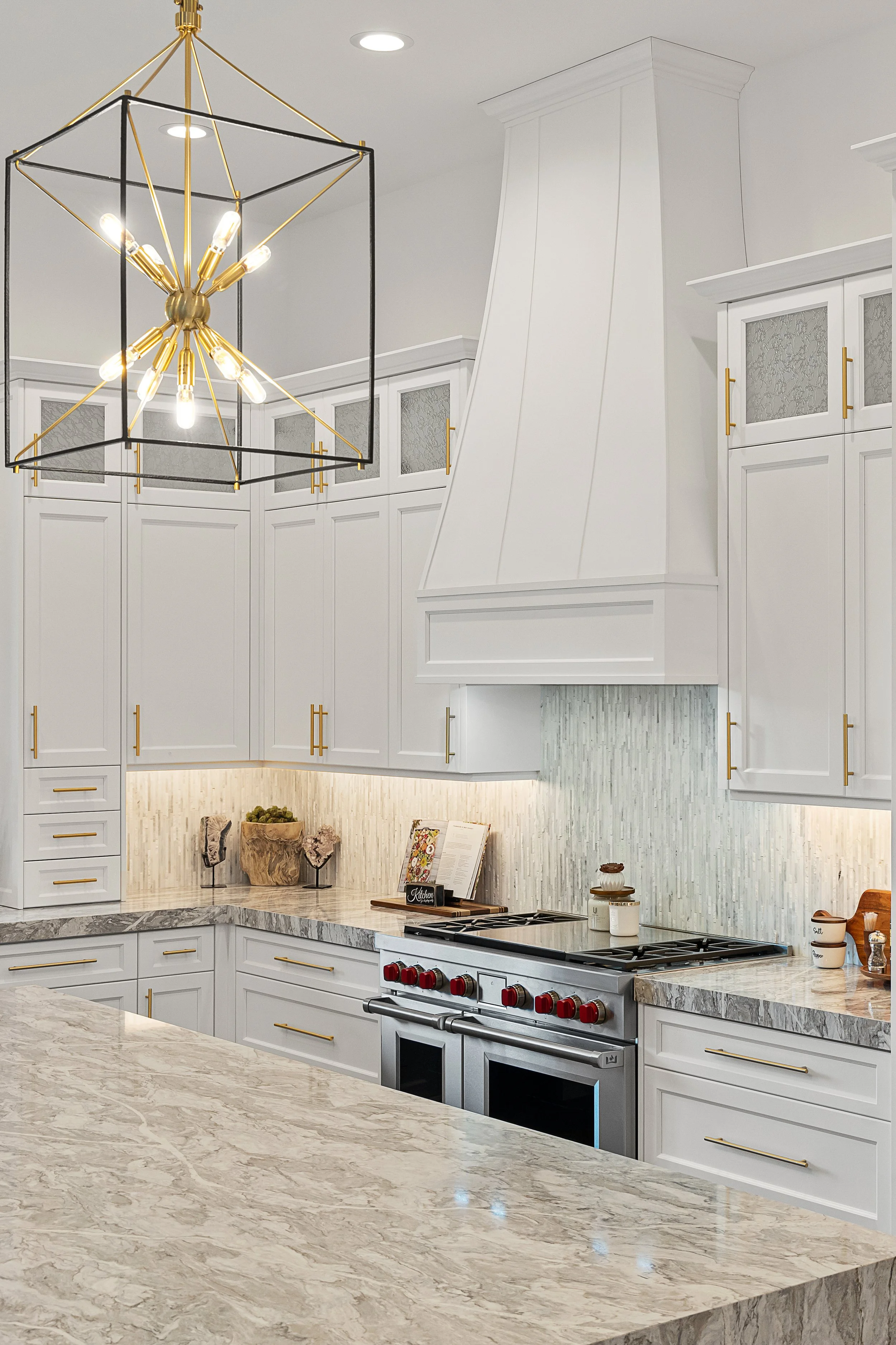 Modern kitchen with white cabinets, gold handles, marble countertops, a gray and white backsplash, a stainless steel stove, and a gold and black geometric chandelier.