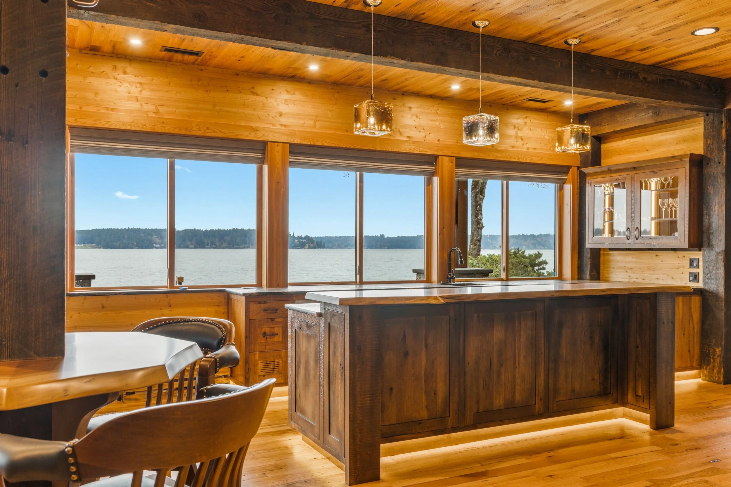 A kitchen with wooden walls, ceiling, and flooring, featuring large windows overlooking a body of water and landscape. There are three pendant lights hanging above a wooden kitchen island, with a sink and faucet. To the left, part of a dining table w