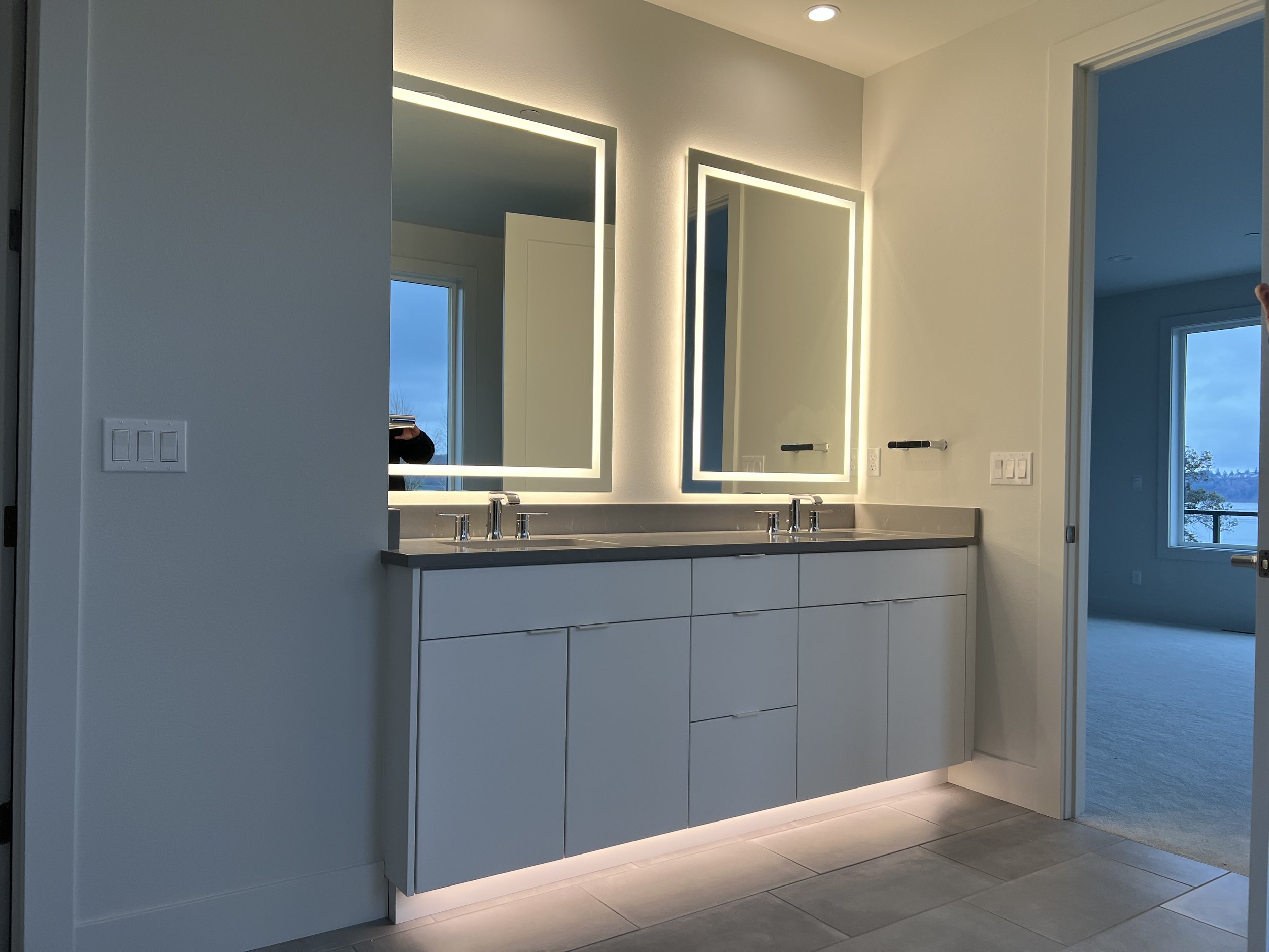 Modern bathroom with double vanity, large illuminated mirrors, and a view of an adjacent bedroom with a window showing a landscape.