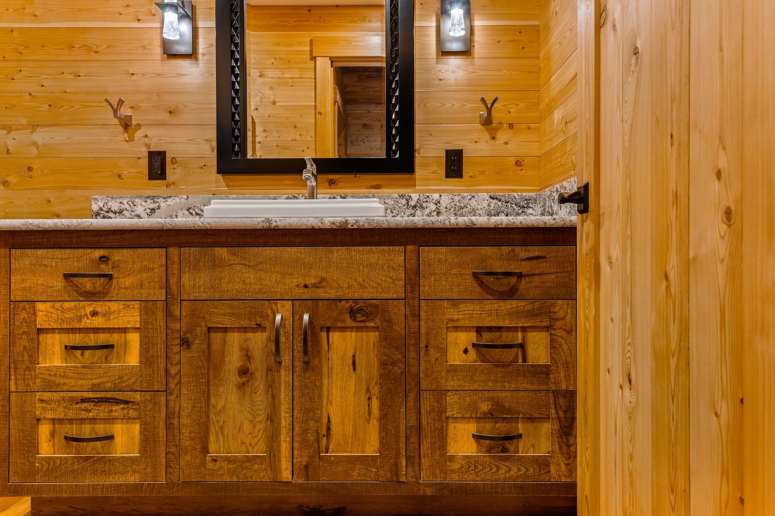 Wooden bathroom vanity with a granite countertop, a white sink, and a mirror above it. The background features wooden walls, a black-framed mirror, and wall-mounted light fixtures.