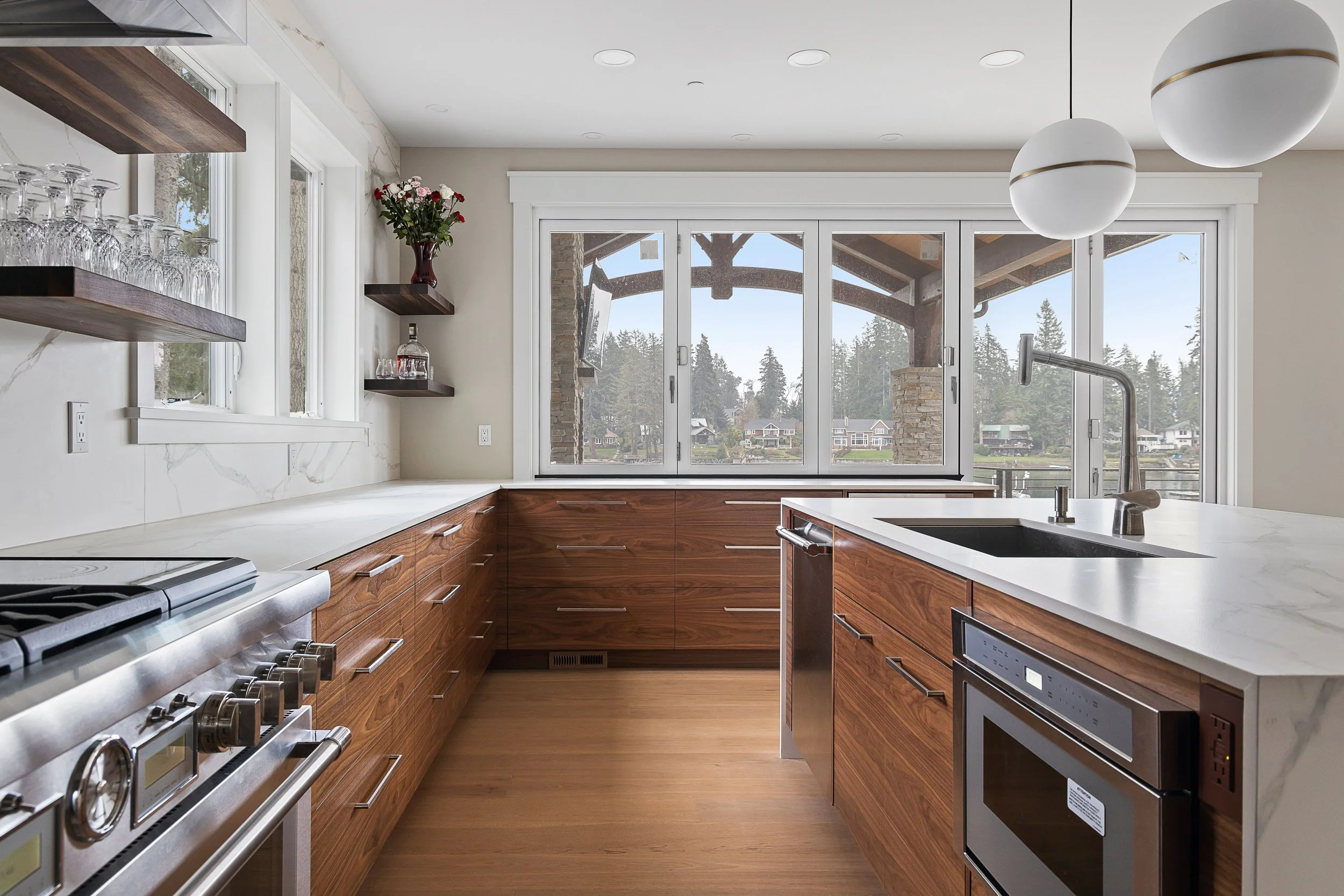 Modern kitchen with wood cabinets, white marble countertops, and large windows overlooking a lake and trees, with pendant lights hanging from the ceiling.