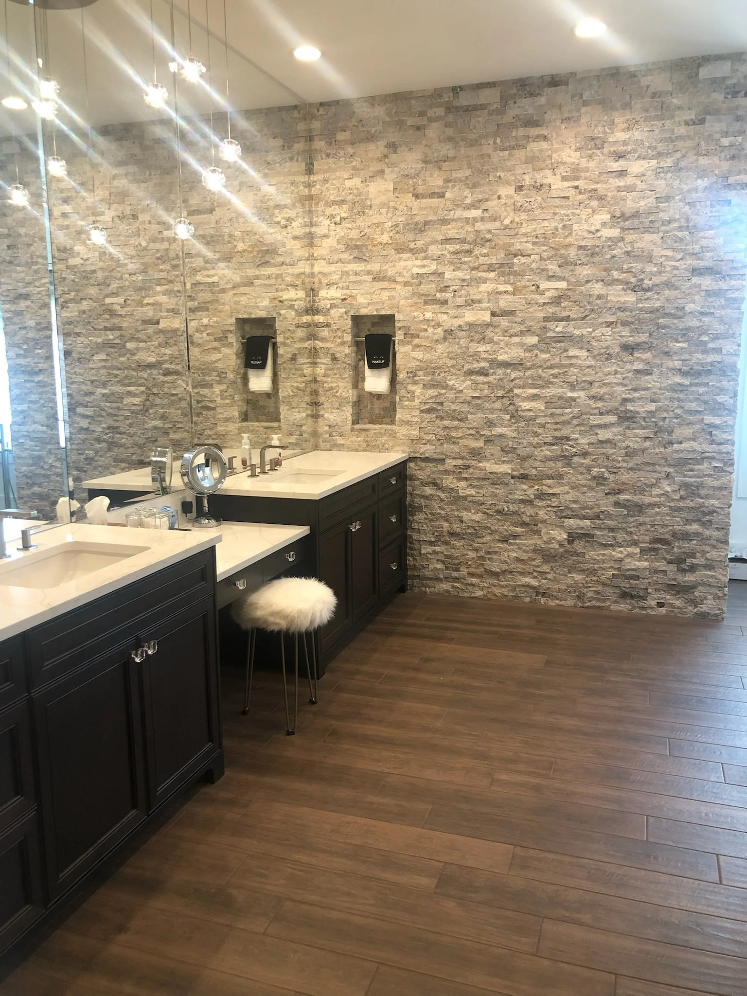 A modern bathroom with a stone accent wall, dark cabinets, marble countertops, and a large mirror. There are two sinks, a stool with a furry cover, and wall-mounted soap dispensers. The lighting is bright with multiple ceiling lights.