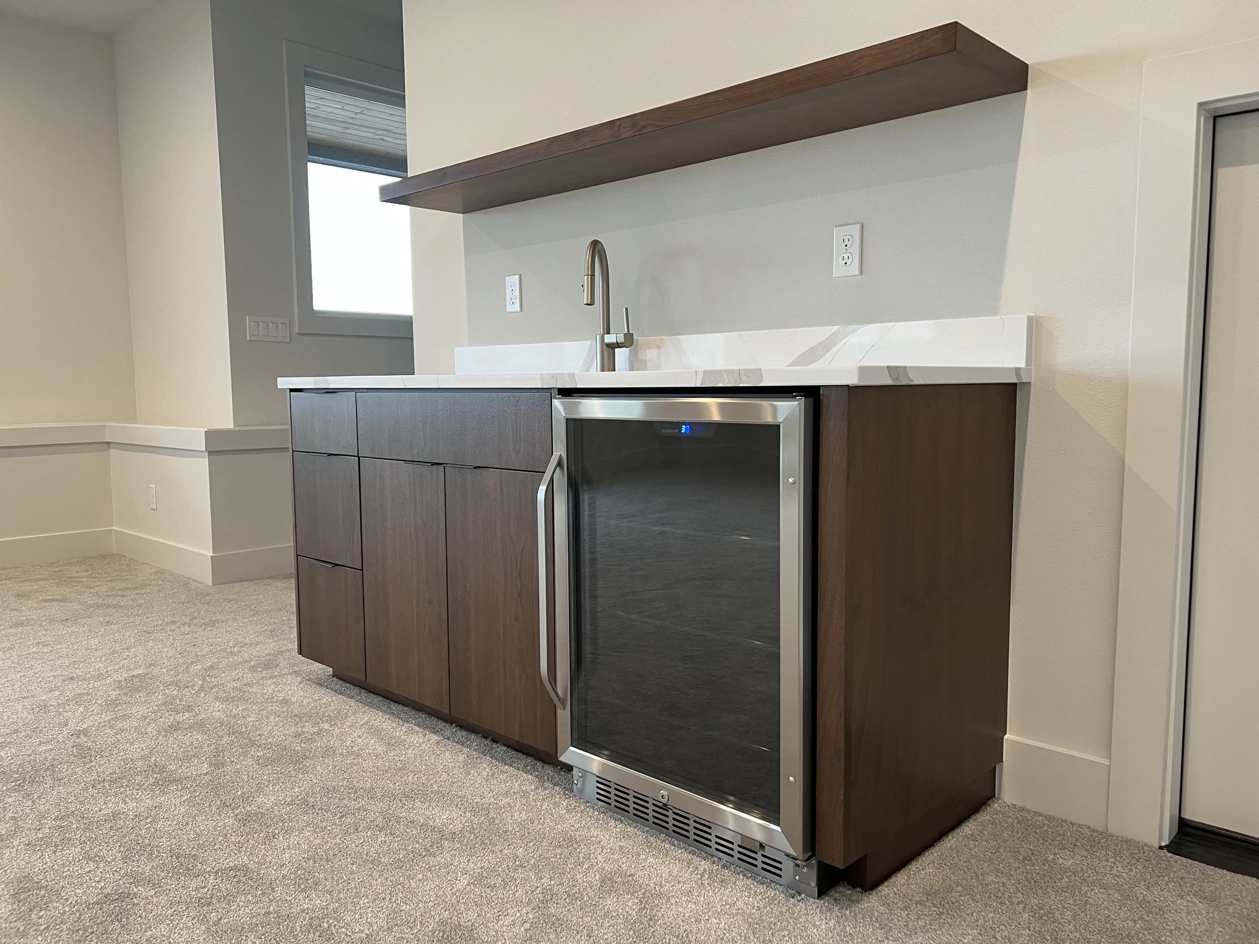 A kitchenette area with a small refrigerator, dark wood cabinets, a white marble countertop, a stainless steel faucet, and a floating wooden shelf on a beige wall.