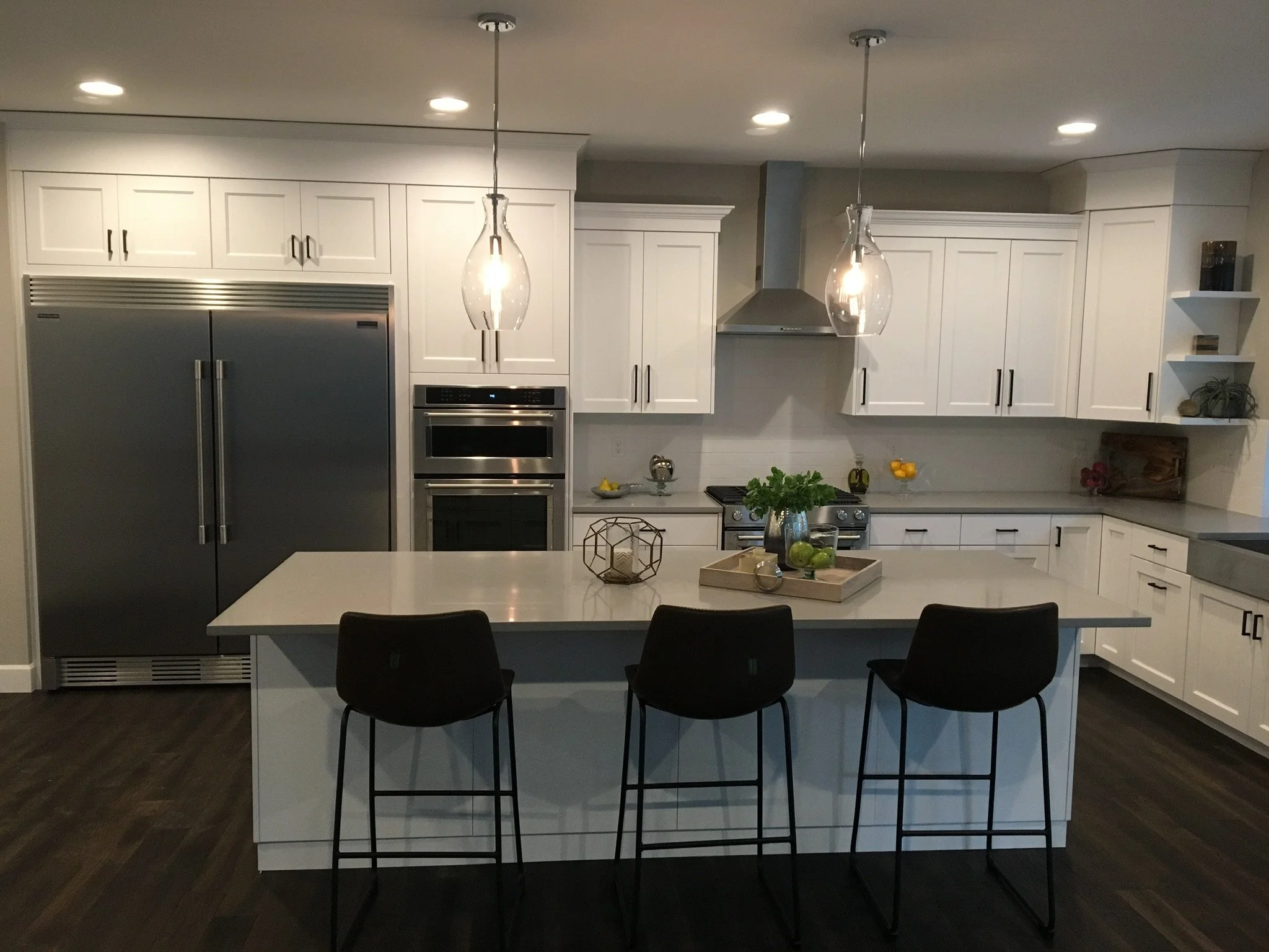 Modern kitchen with white cabinets, stainless steel refrigerator and oven, dark wood floor, kitchen island with three black chairs, pendant lights, and decorative items including a plant and fruits.