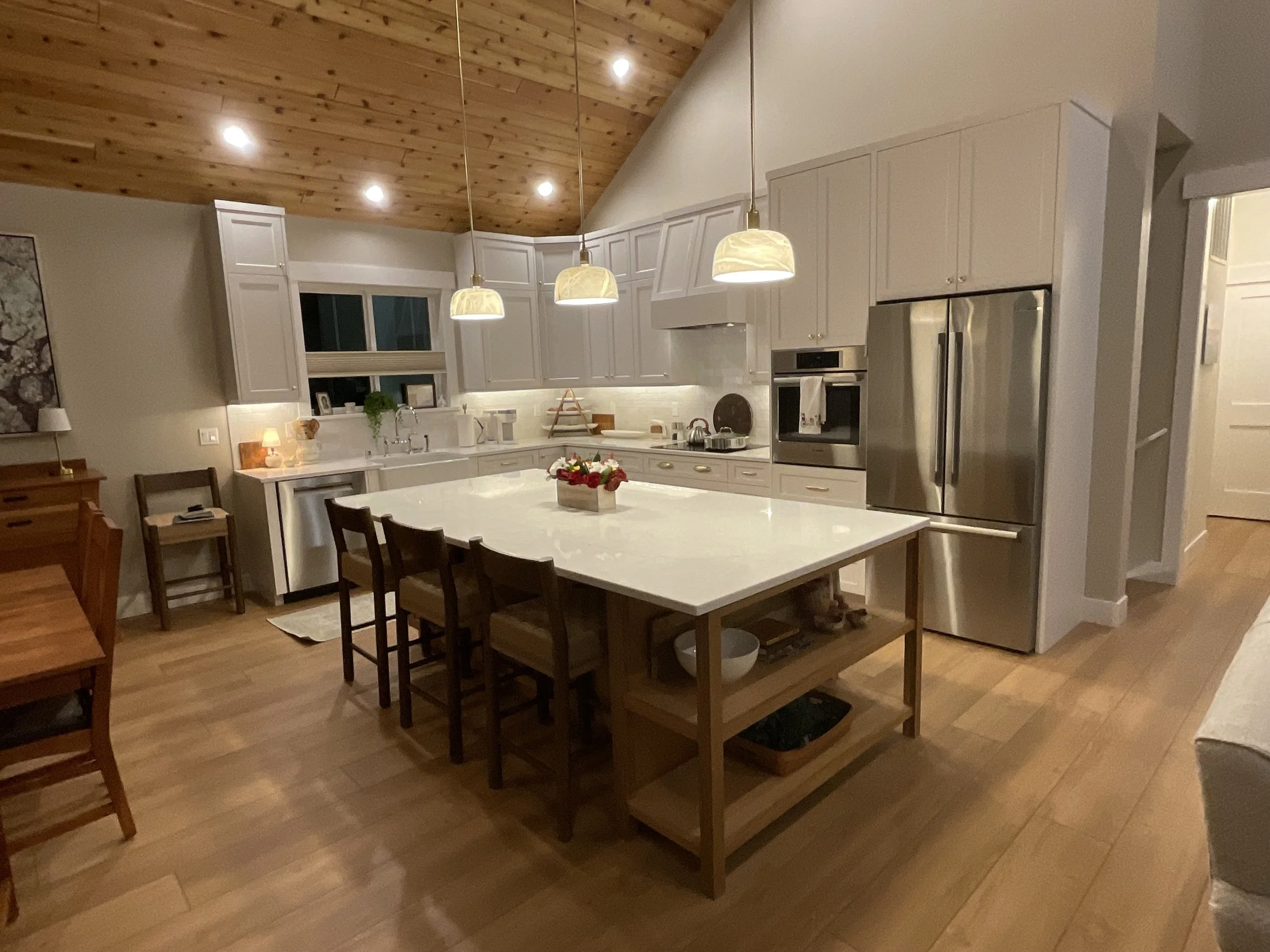 Kitchen with white cabinets, stainless steel refrigerator, and a large island with a white countertop and wooden base, illuminated by hanging pendant lights with cream-colored shades, and warm wood flooring.