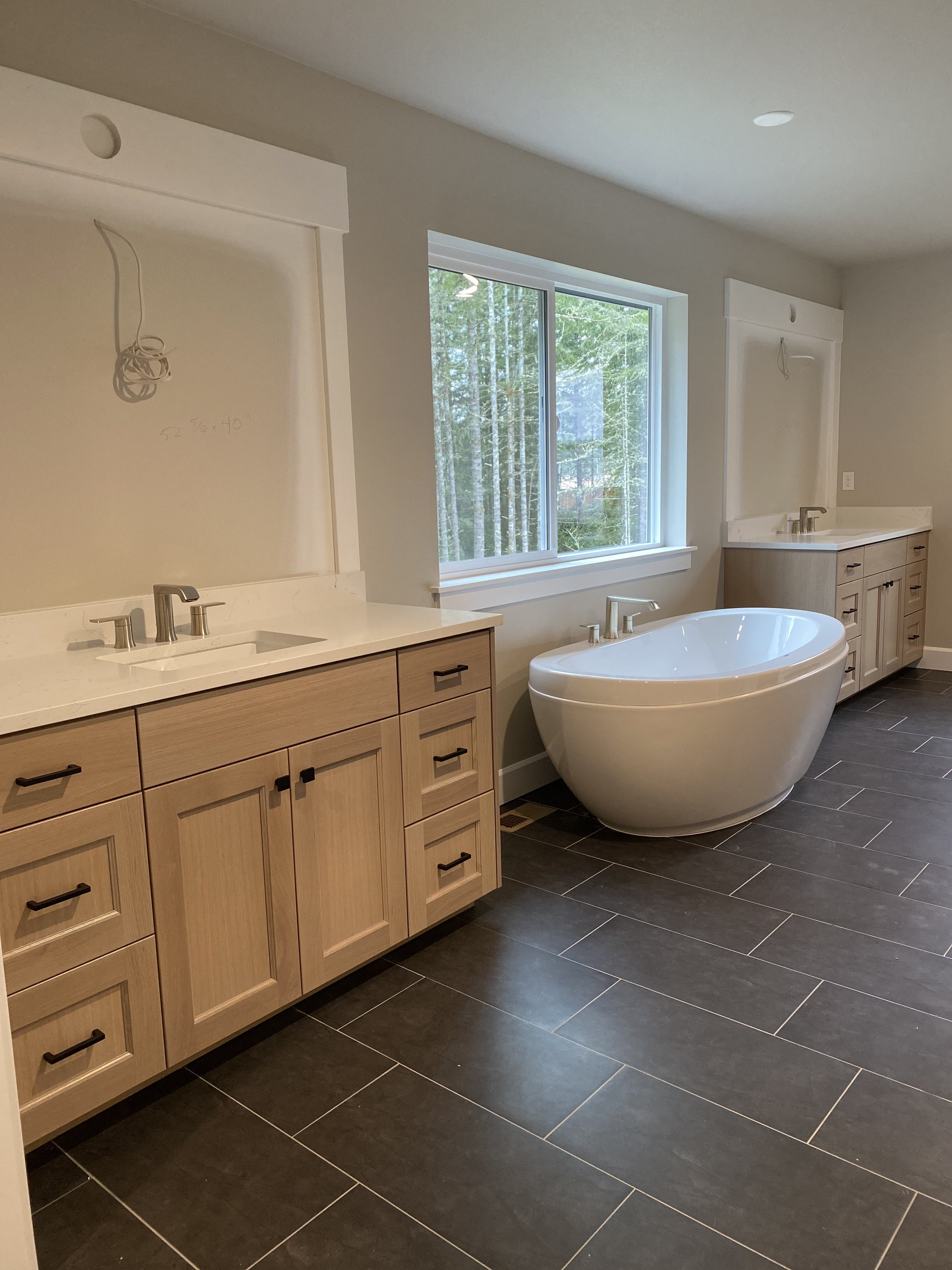 A modern bathroom with light wood cabinetry, a white freestanding bathtub, and a large window with trees outside.