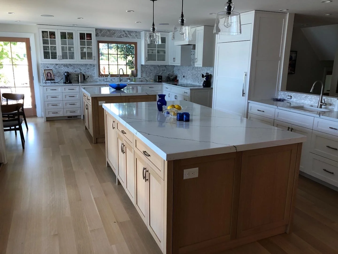Modern kitchen with island, white cabinets, light wood flooring, and large windows, featuring blue and yellow decor and pendant lights.