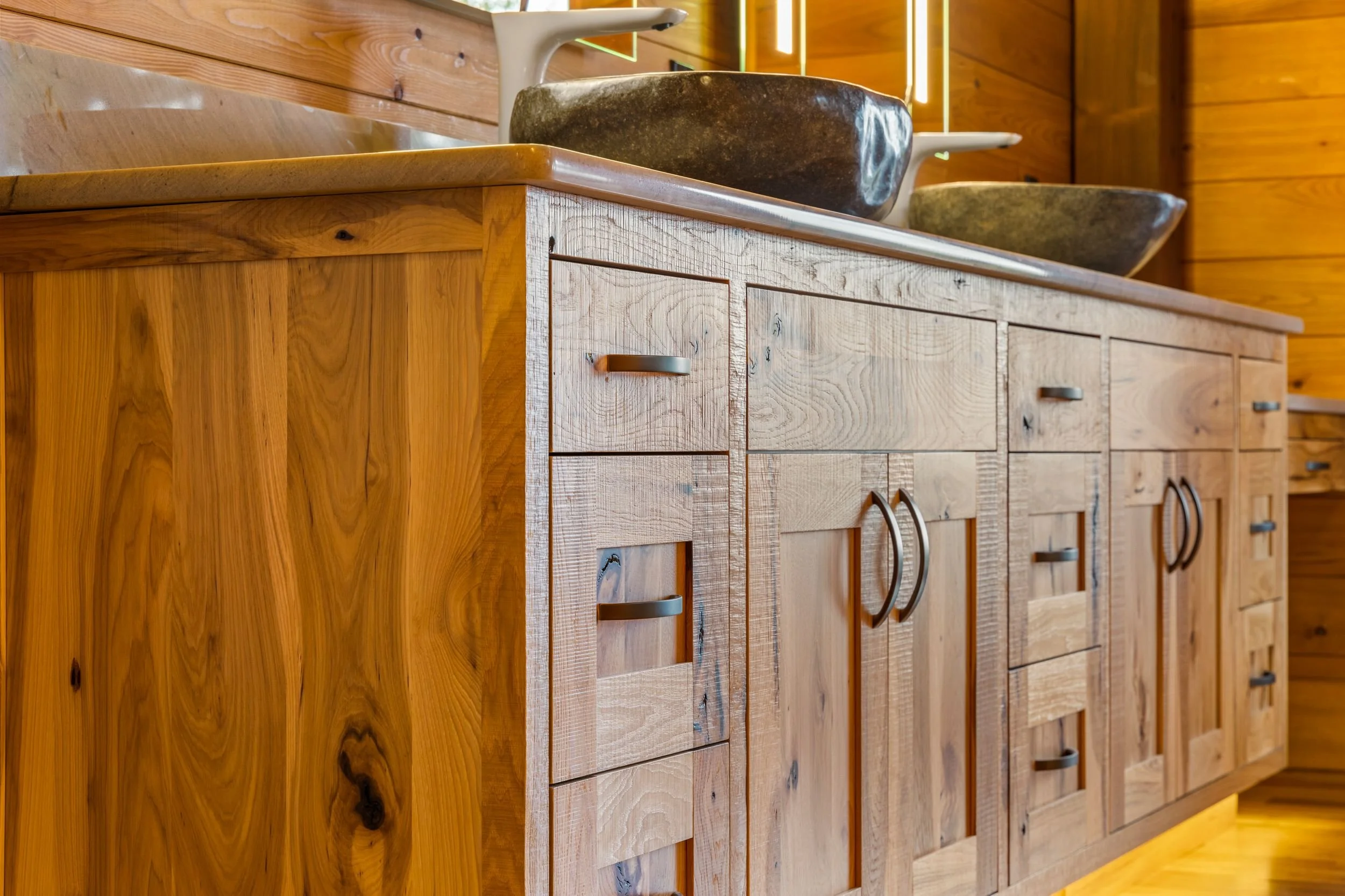 A wooden bathroom vanity with multiple drawers and cabinets, topped with a light-colored countertop, featuring two stone sinks and white faucets.
