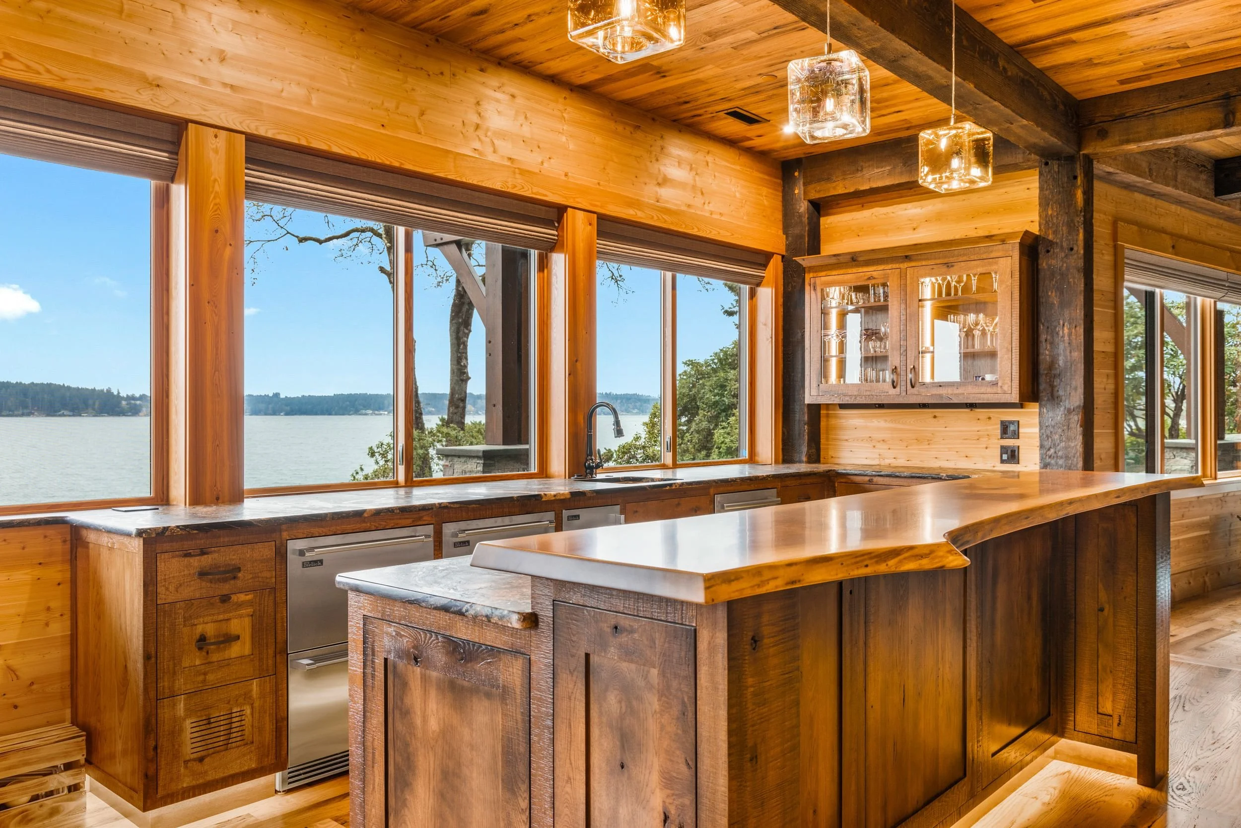 Rustic kitchen with wooden cabinets and countertops, large windows overlooking a lake, and modern pendant lights hanging from a wooden ceiling.