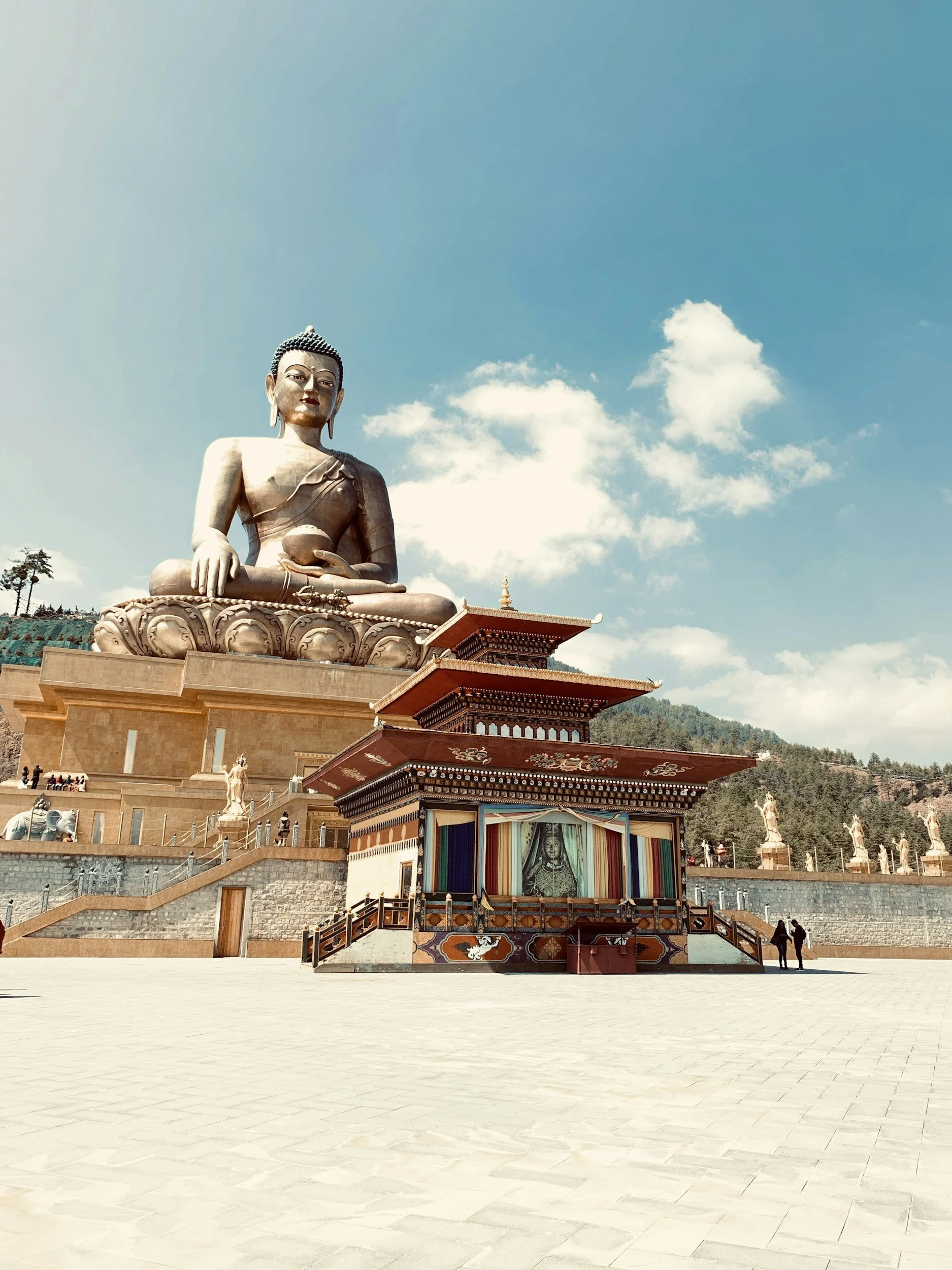 Large seated bronze Buddha statue overlooking a traditional Tibetan-style building with colorful fabric drapes, against a background of mountains and blue sky.