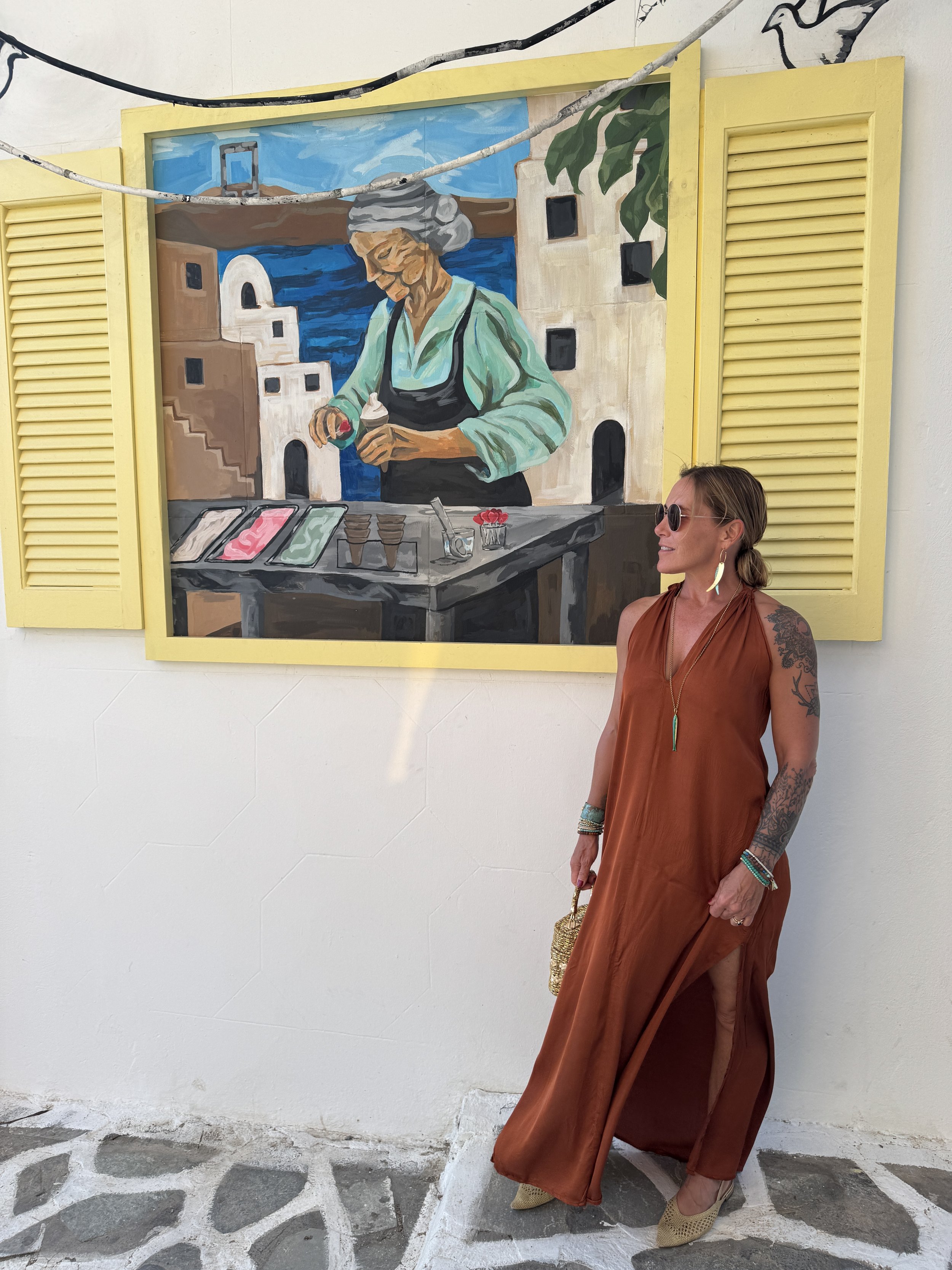 Woman in a brown dress with tattoos, sunglasses, and jewelry standing in front of a painting of an older woman making ice cream on a seaside terrace with yellow shutters.
