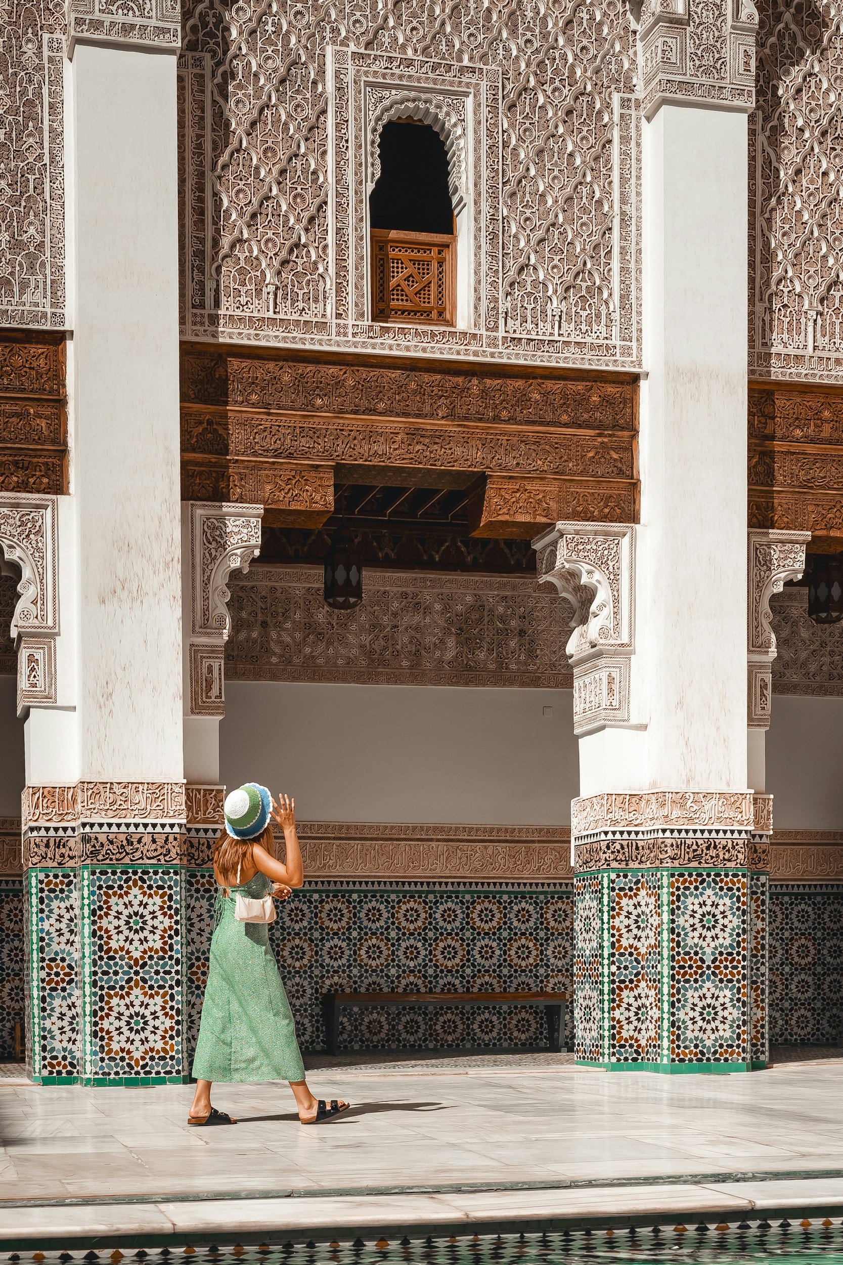 A woman in a green dress and large hat looks up at detailed Moroccan-style architecture with white walls, intricate carvings, and patterned tilework.
