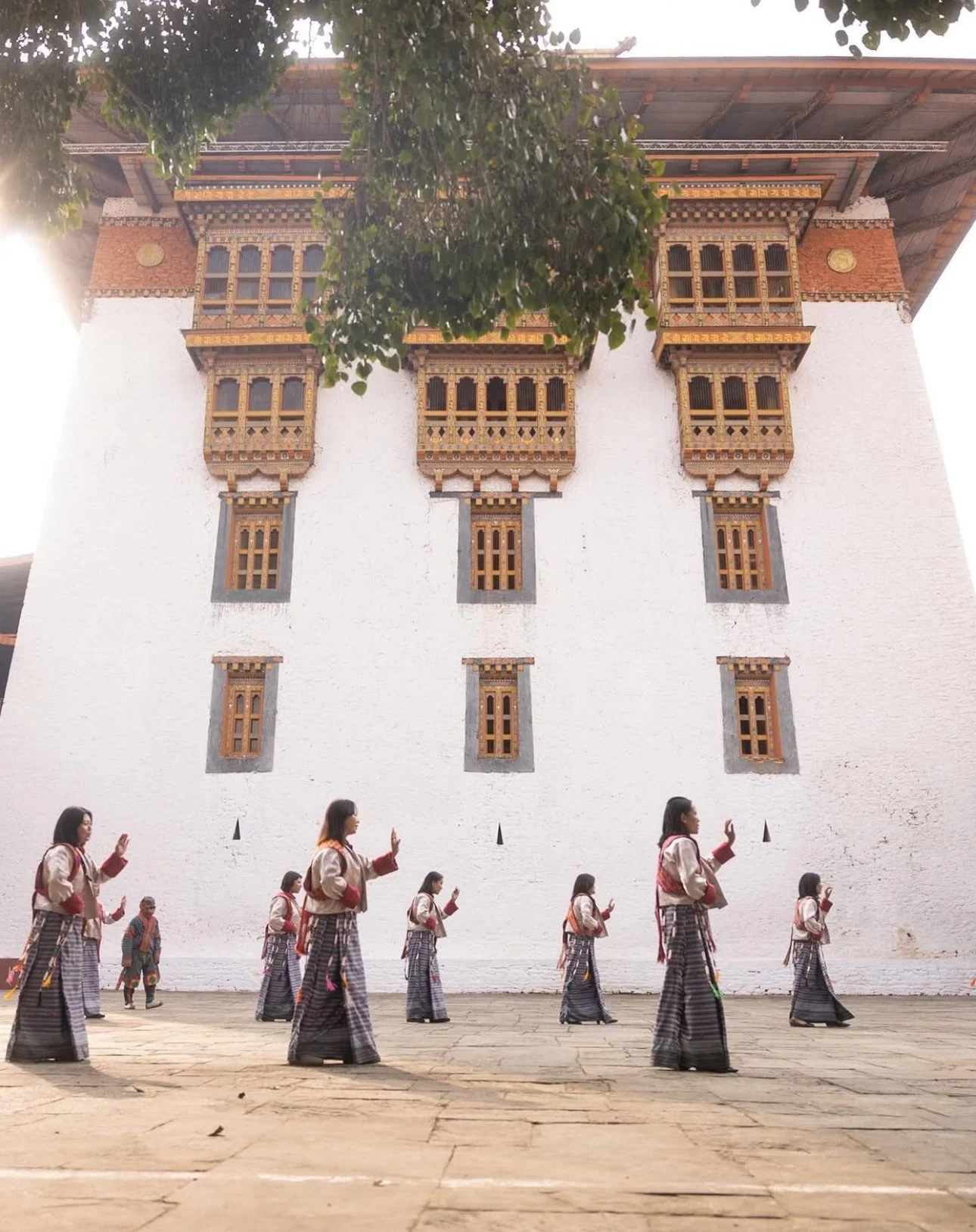 Group of people performing a traditional dance in front of a white wall with ornate wooden windows, with a large tree partially visible at the top.
