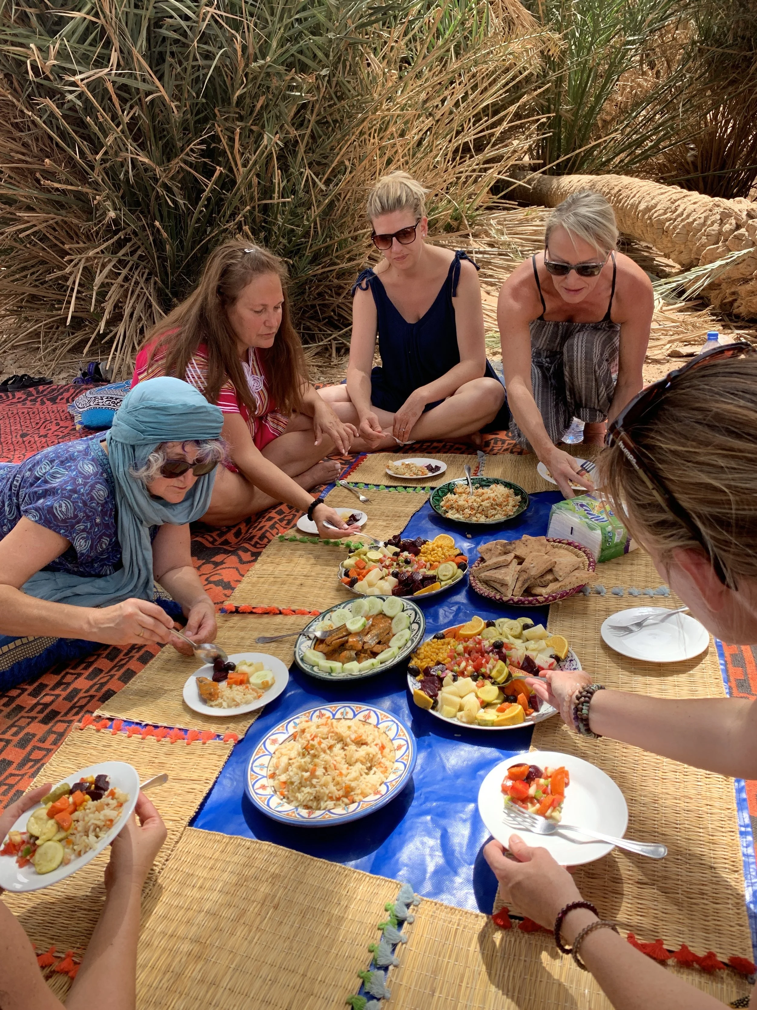 Group of women having a beach picnic with food on a mat in front of desert plants.