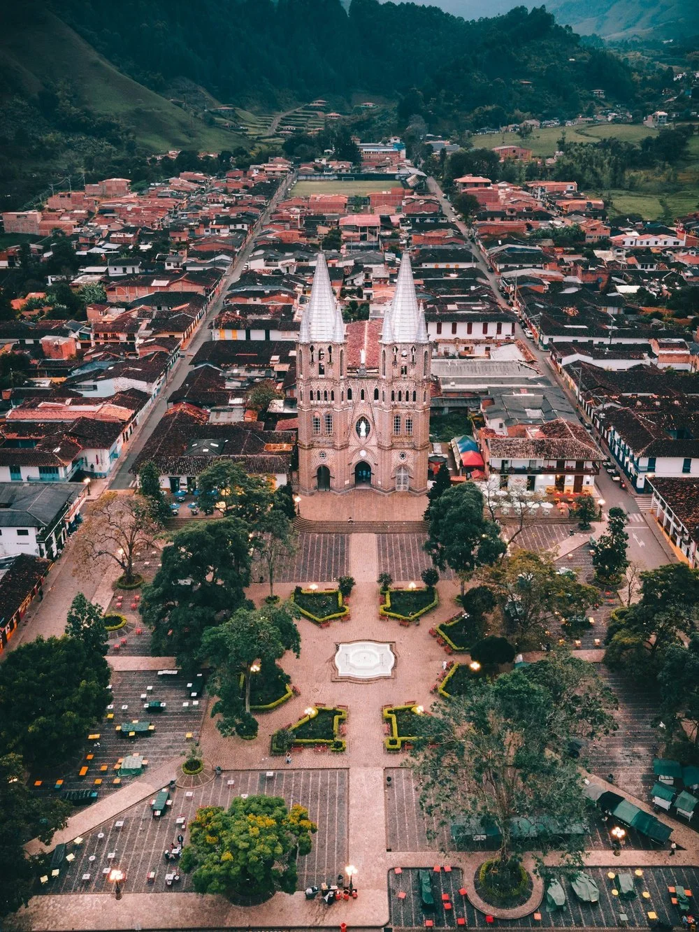 An aerial view of a historic church with twin spires located in a town square, surrounded by townhouses and green hills in the background.