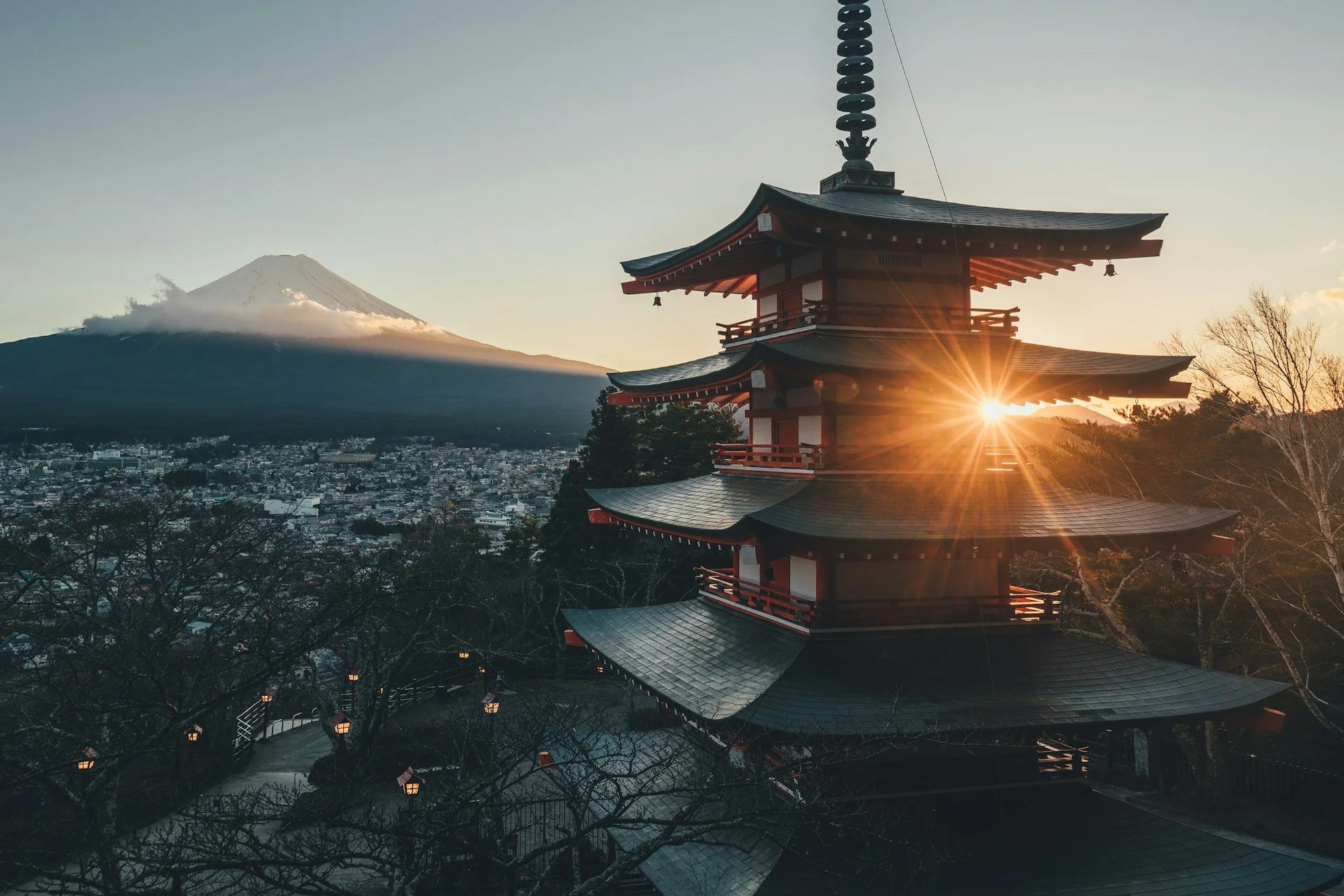 A traditional Japanese pagoda with multiple tiers, situated in a scenic area during sunset, with Mount Fuji in the background and the sun creating a starburst effect.