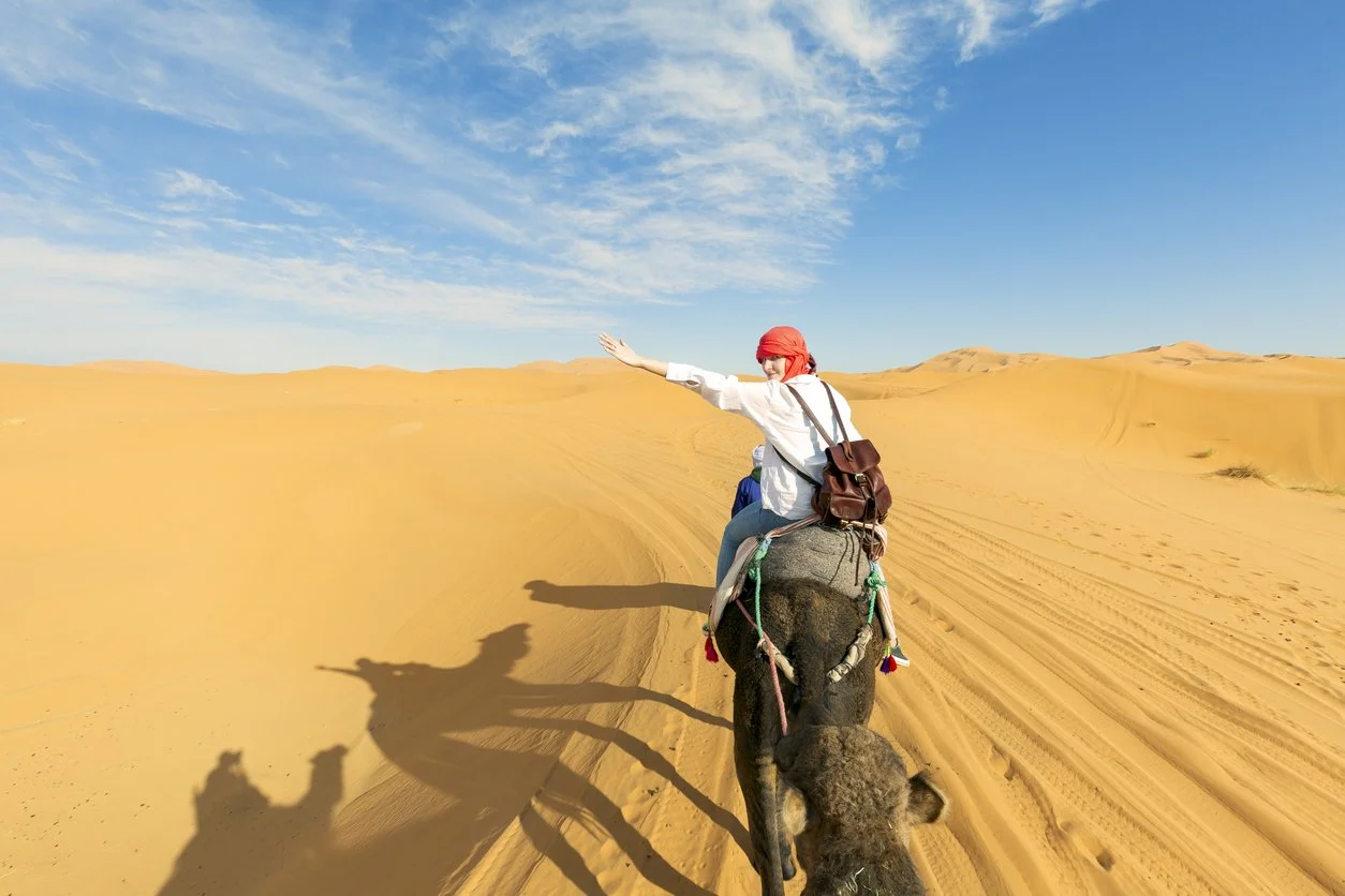 Person riding an elephant through a desert with sand dunes and a blue sky.