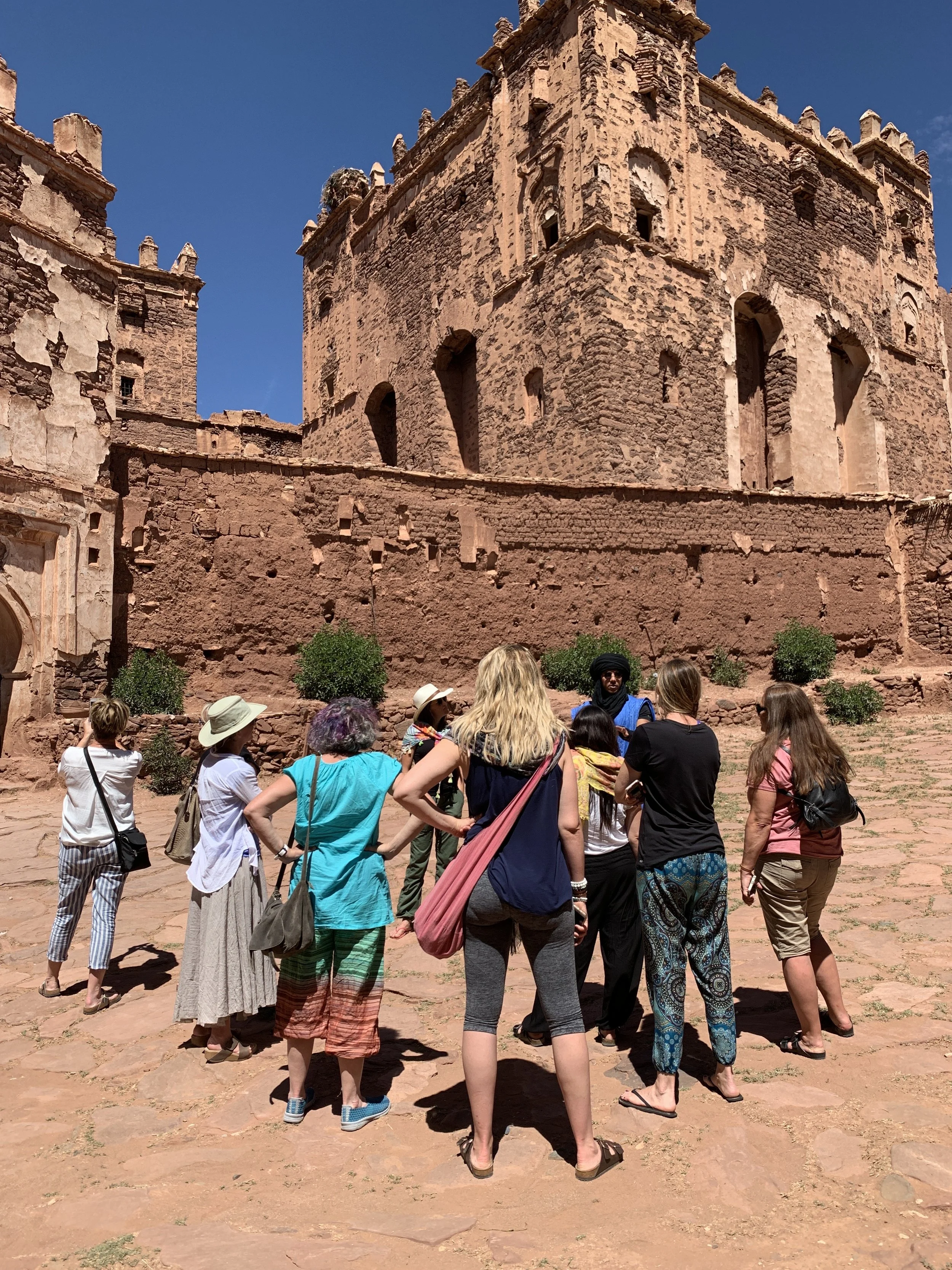 Tour group listening to guide in front of red sandstone castle ruins with blue sky.