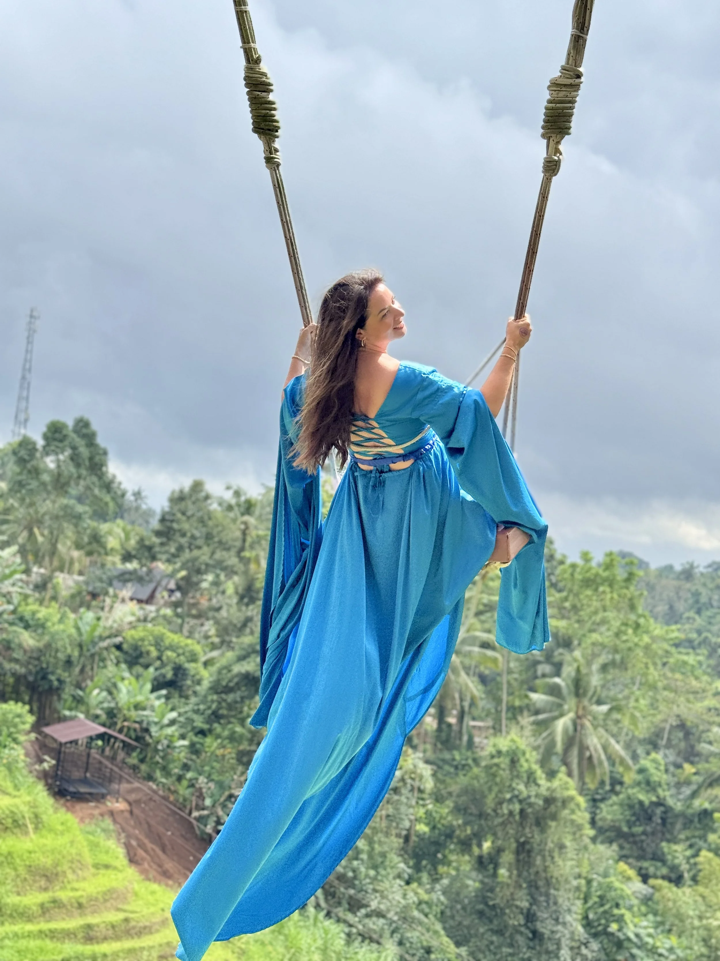 A woman in a flowing blue dress sitting on a swing high above green tropical foliage and rice terraces, with a cloudy sky in the background.
