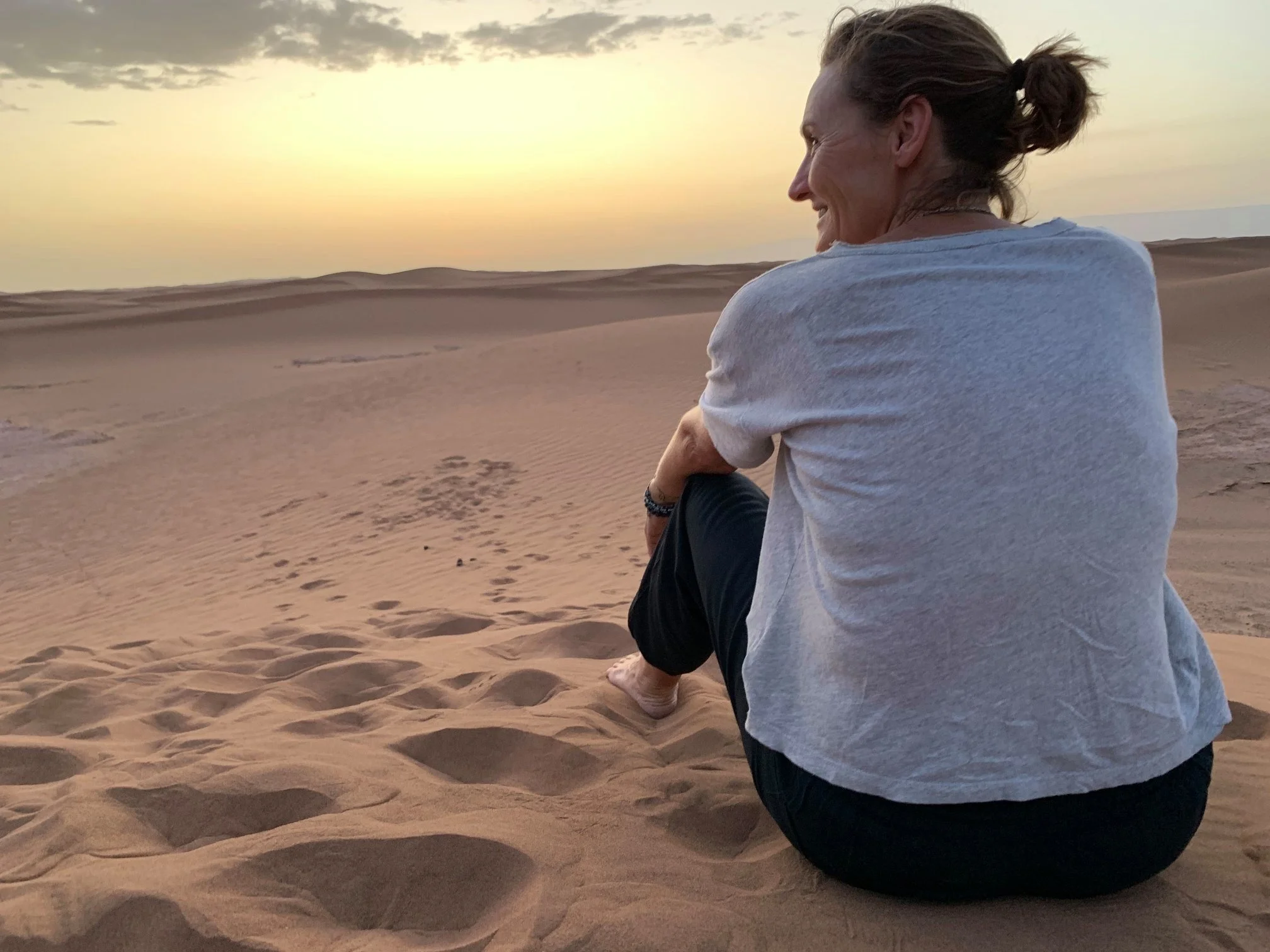 Woman sitting on sand dunes during sunset, smiling and enjoying the view.