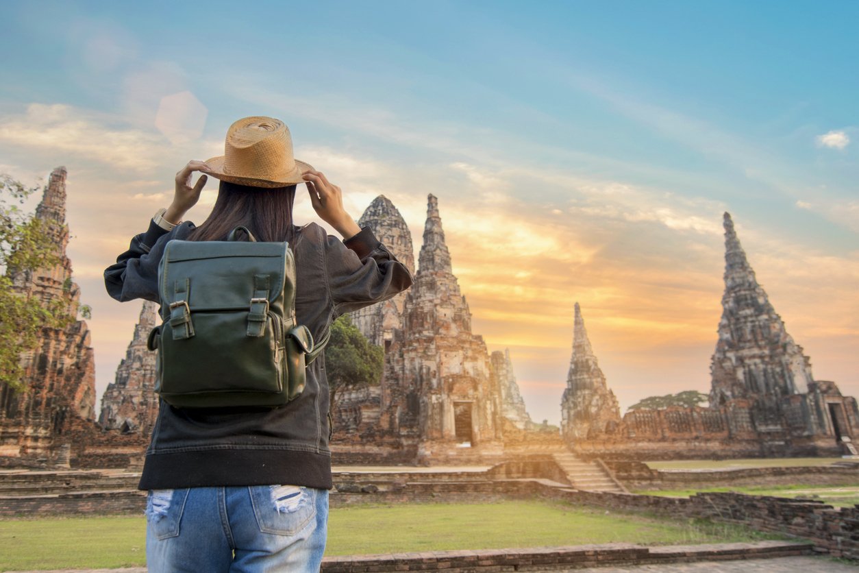 A woman with a backpack and a sun hat looking at ancient temple ruins during sunset.