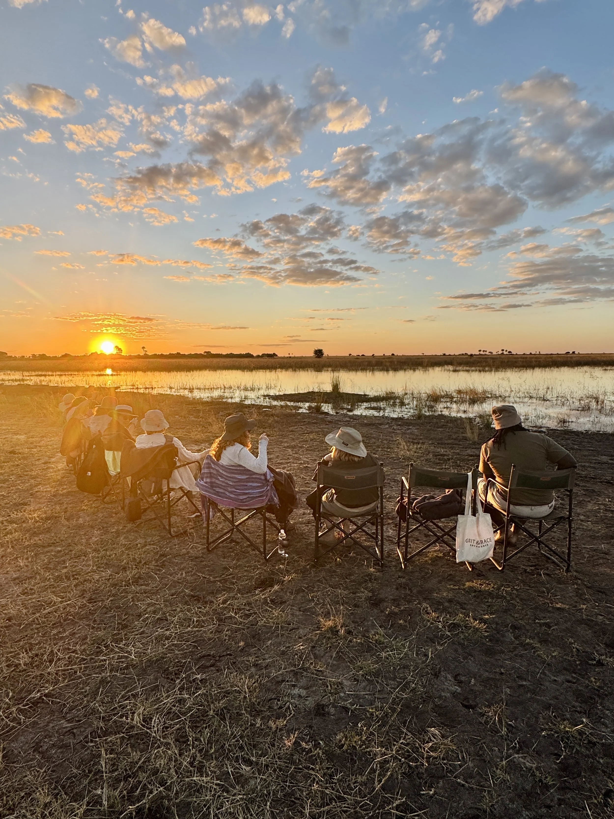 People sitting in chairs watching a sunset over a marshland, some wearing hats, during a group outdoor gathering.