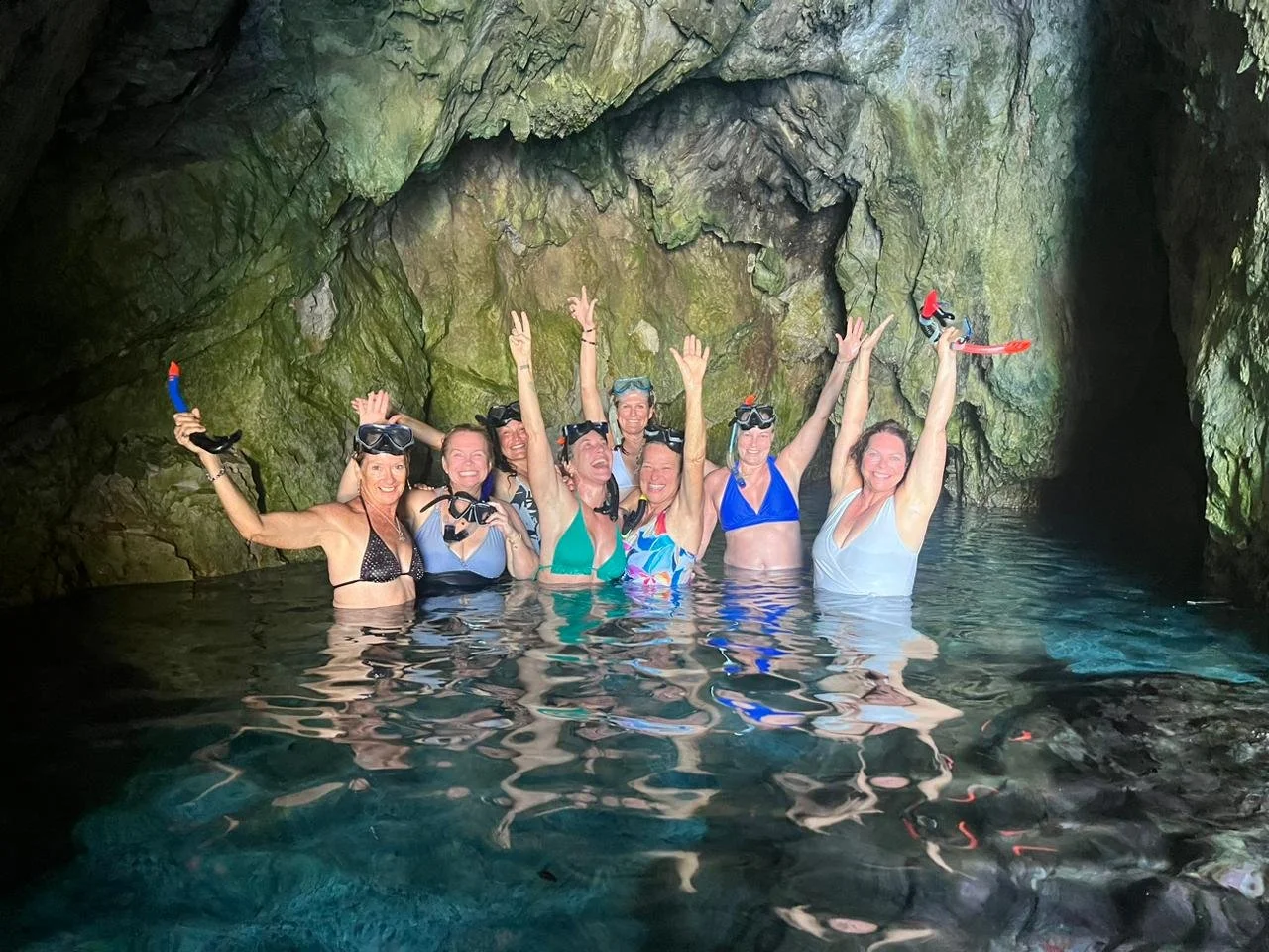 Group of women in swimsuits and snorkeling gear celebrating inside a cave with water up to their chests, posing with arms raised.