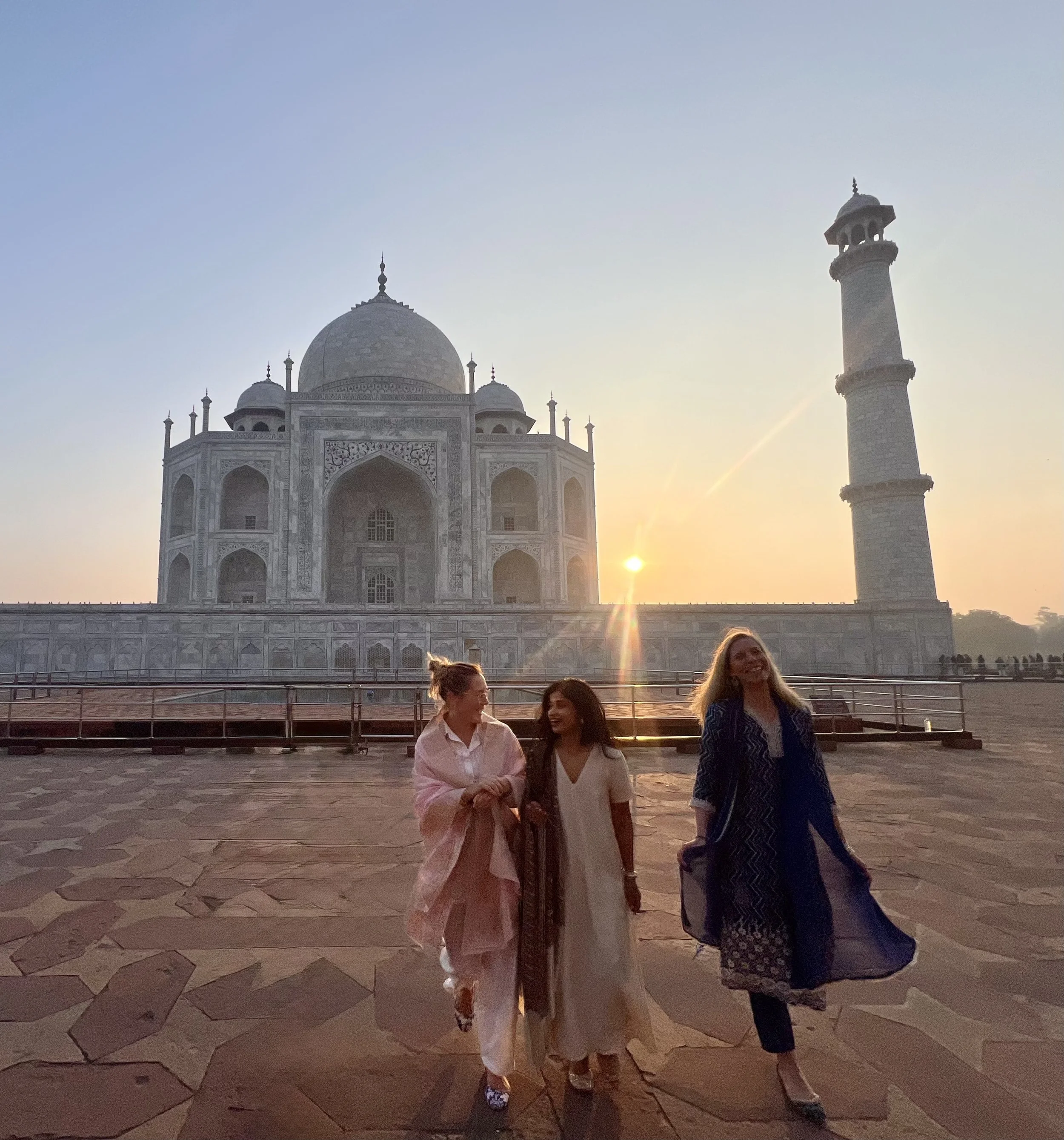 Three women walking near the Taj Mahal at sunrise, with the iconic white marble mausoleum and its minaret in the background.