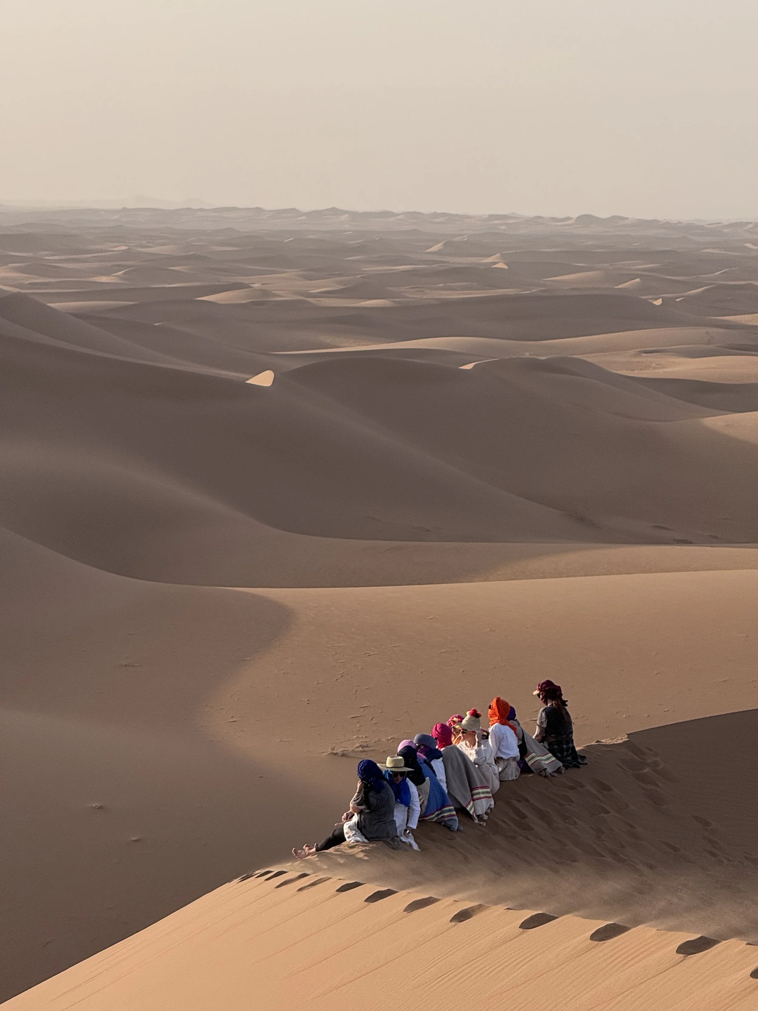 Group of people sitting on the sand dunes in the desert, wearing colorful clothing and head coverings.