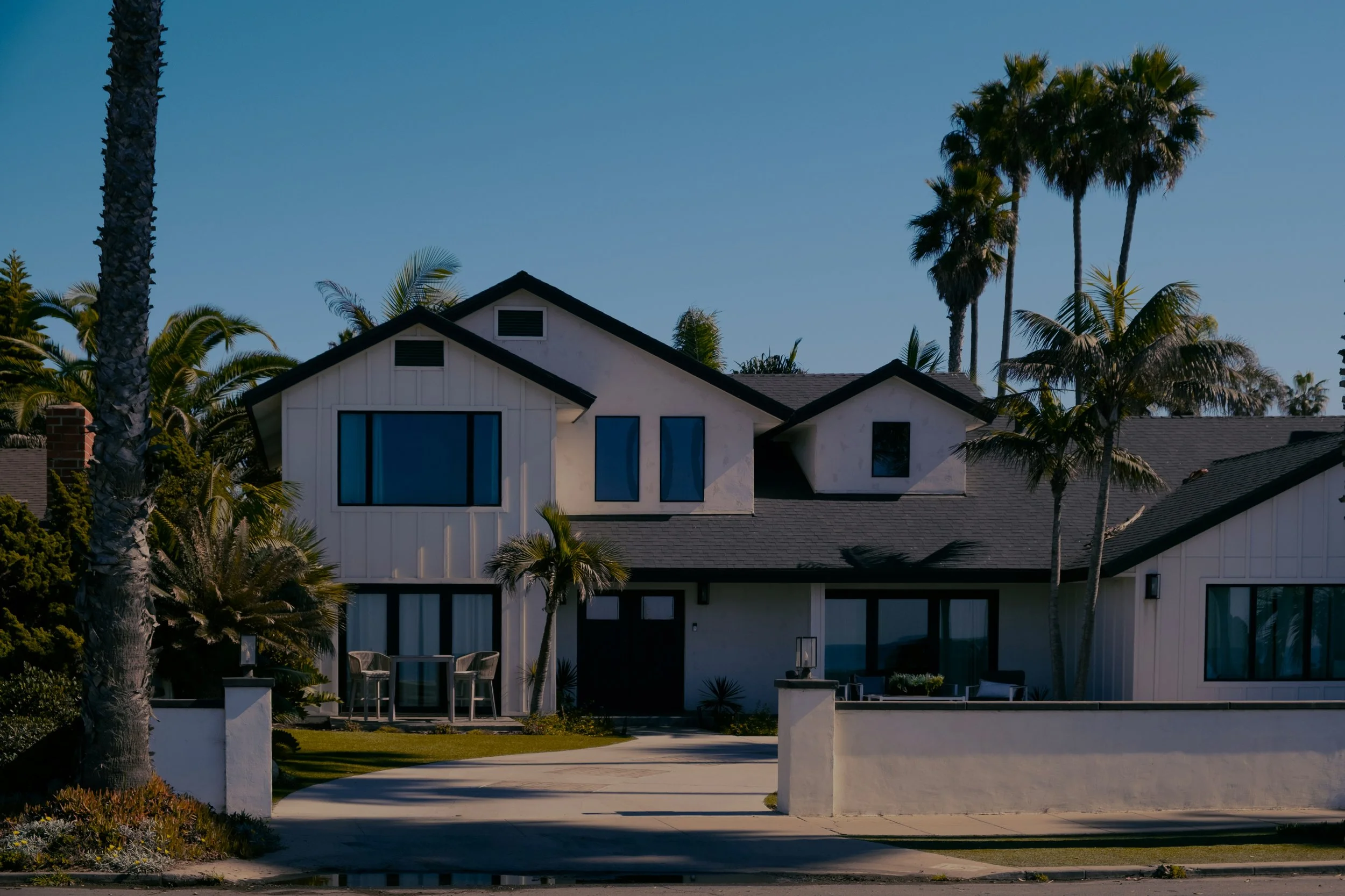 Modern white house with large windows situated among palm trees and a clear blue sky.