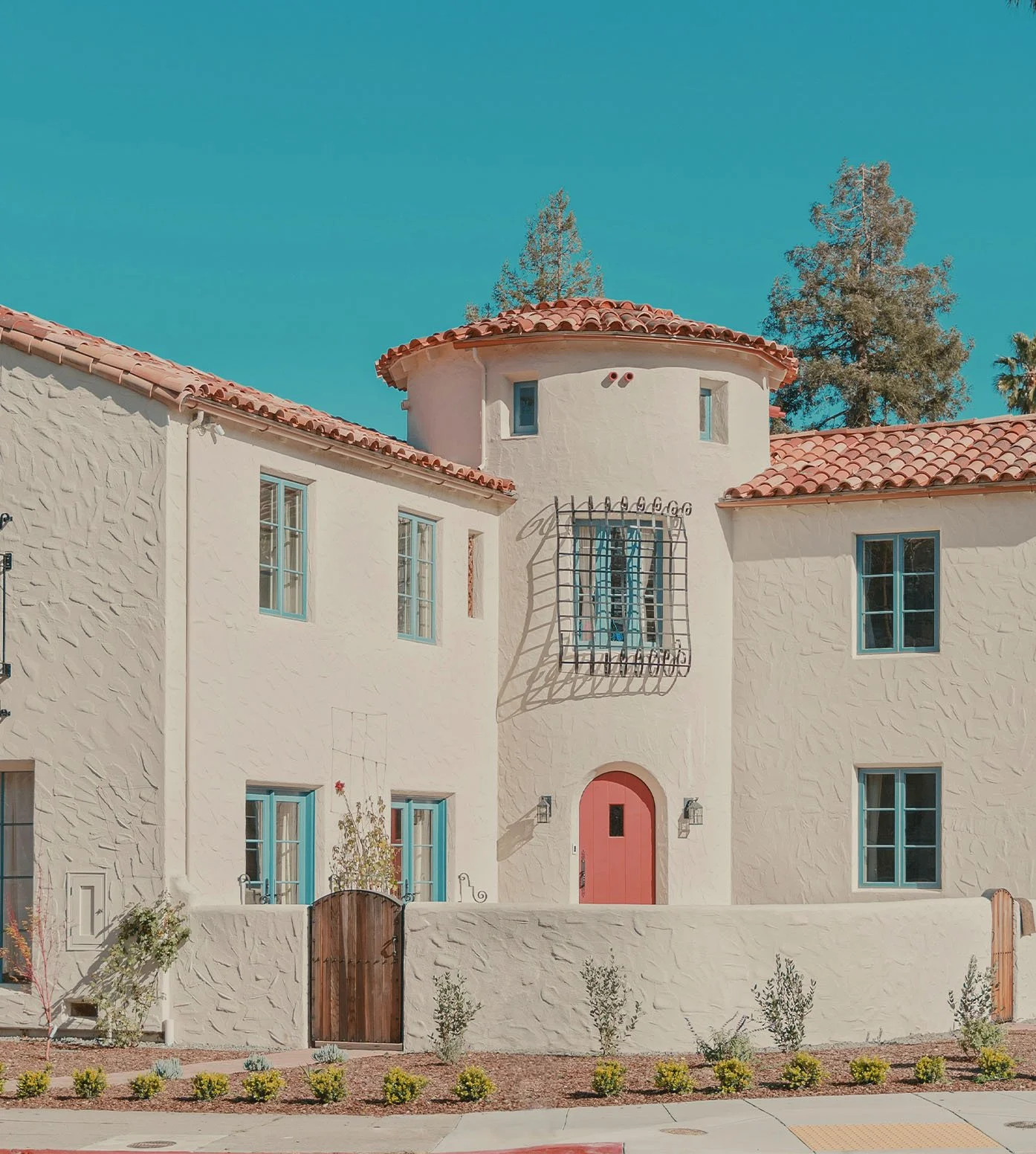 A Mediterranean-style house with white stucco walls, red tile roof, pink arched front door, and blue window frames, surrounded by a small landscaped front yard with bushes and a wooden gate.