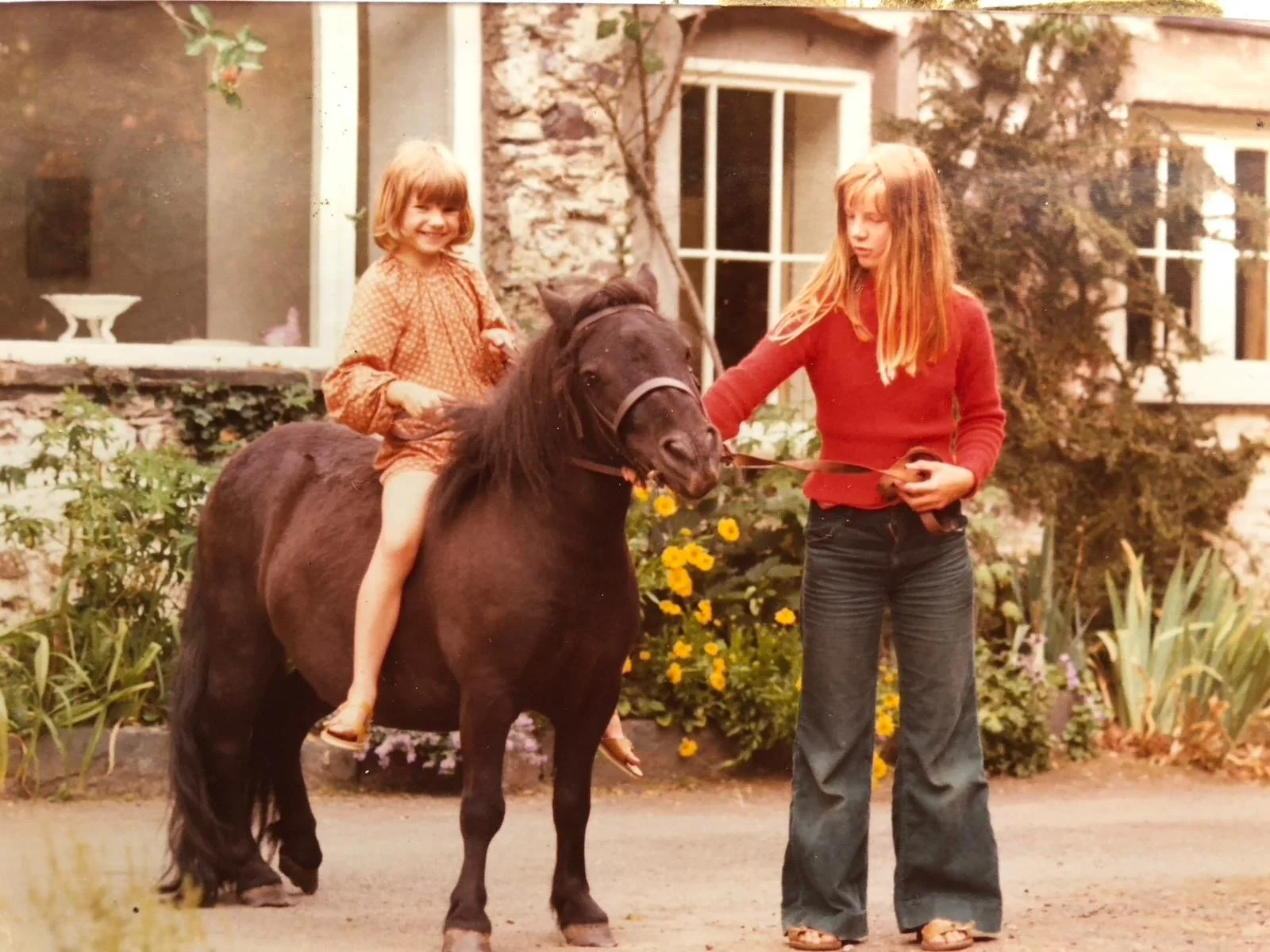 A young girl with blonde hair sitting on a small black pony in a garden, smiling. An older girl with red hair holding the pony's bridle, standing on the ground. There are yellow flowers and green foliage in the background, along with a house with lar