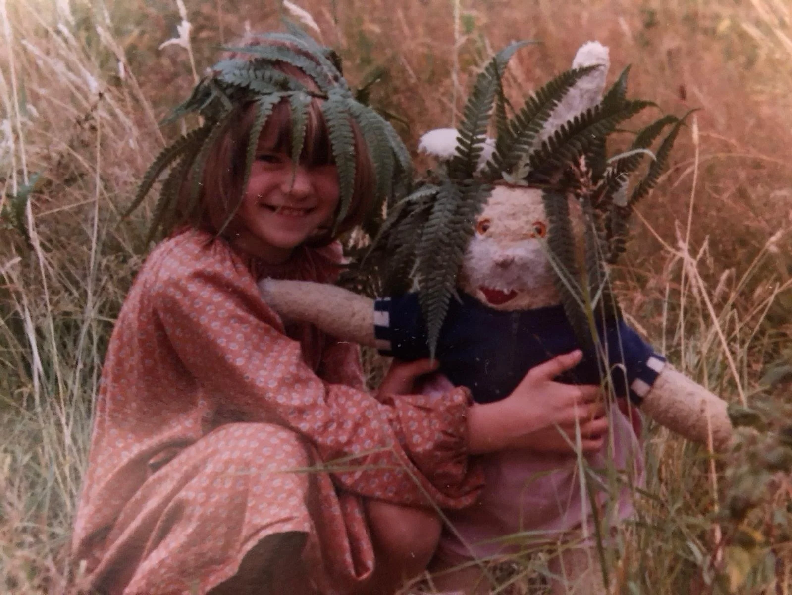 Child with a face mask and oversized fern hat holding a stuffed animal with a fern hat, in a grassy field.